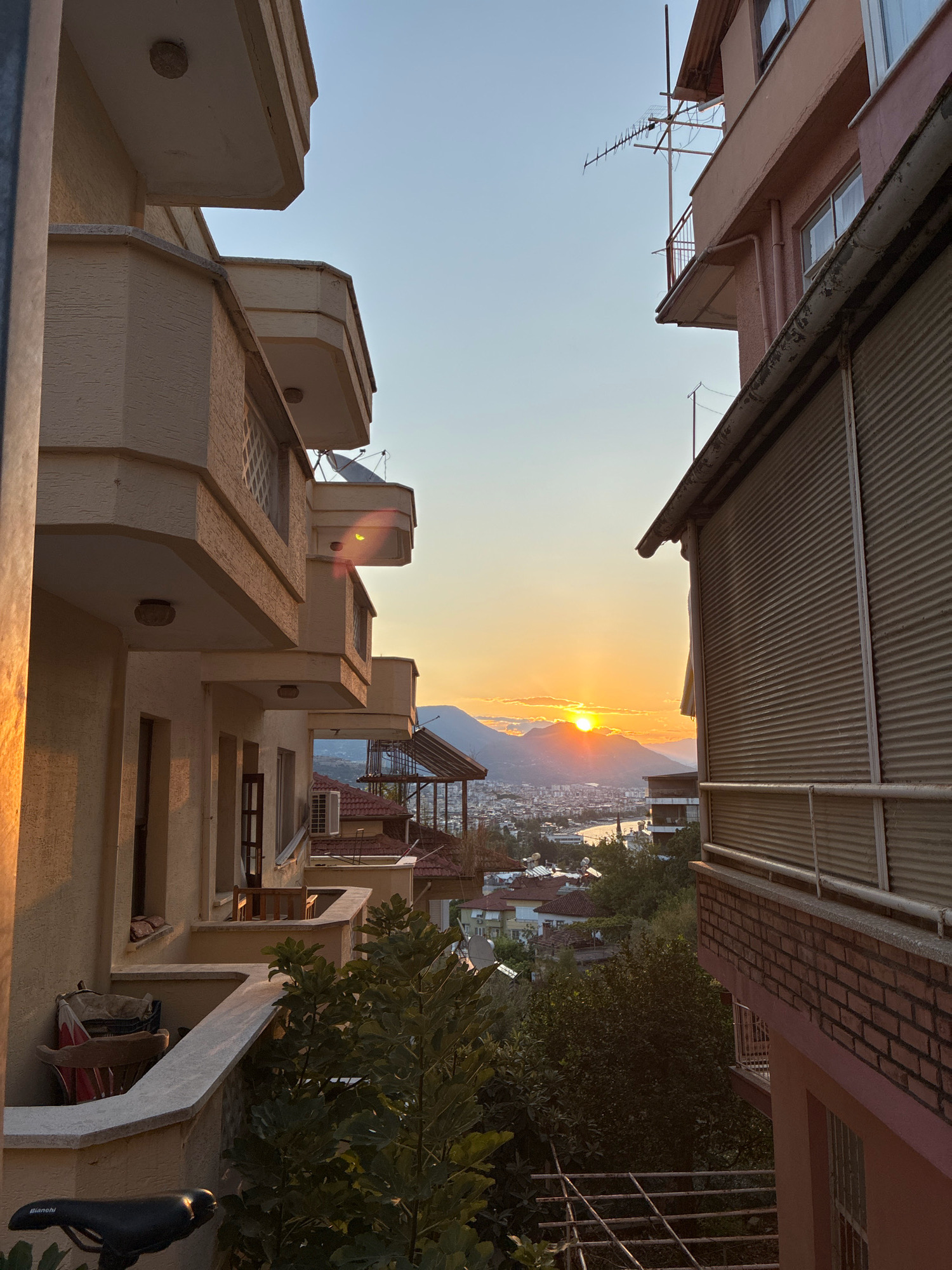 This is a photograph taken from a residential building overlooking the city of Alanya, Turkey, during sunset. The sun is low on the horizon, casting a warm golden glow over the scene and partially obscured by mountains in the distance. In the foreground, a pale yellow apartment building with multiple balconies is visible on the left. The balconies have angular, concrete overhangs and some have satellite dishes. On the right, a portion of a pink apartment building is seen, featuring a metal roller shutter and a brick base. Between the buildings, the cityscape of Alanya unfolds, with a body of water, likely the Mediterranean Sea, visible in the distance. Numerous residential buildings, some with red-tiled roofs, are scattered across the hillside, interspersed with green trees. The sky is a clear gradient of pale blue to orange, indicating the time of day. The composition is framed by the architecture of the apartment buildings, creating a sense of looking out from a private residence onto the broader urban landscape. There are no people visible in the image.