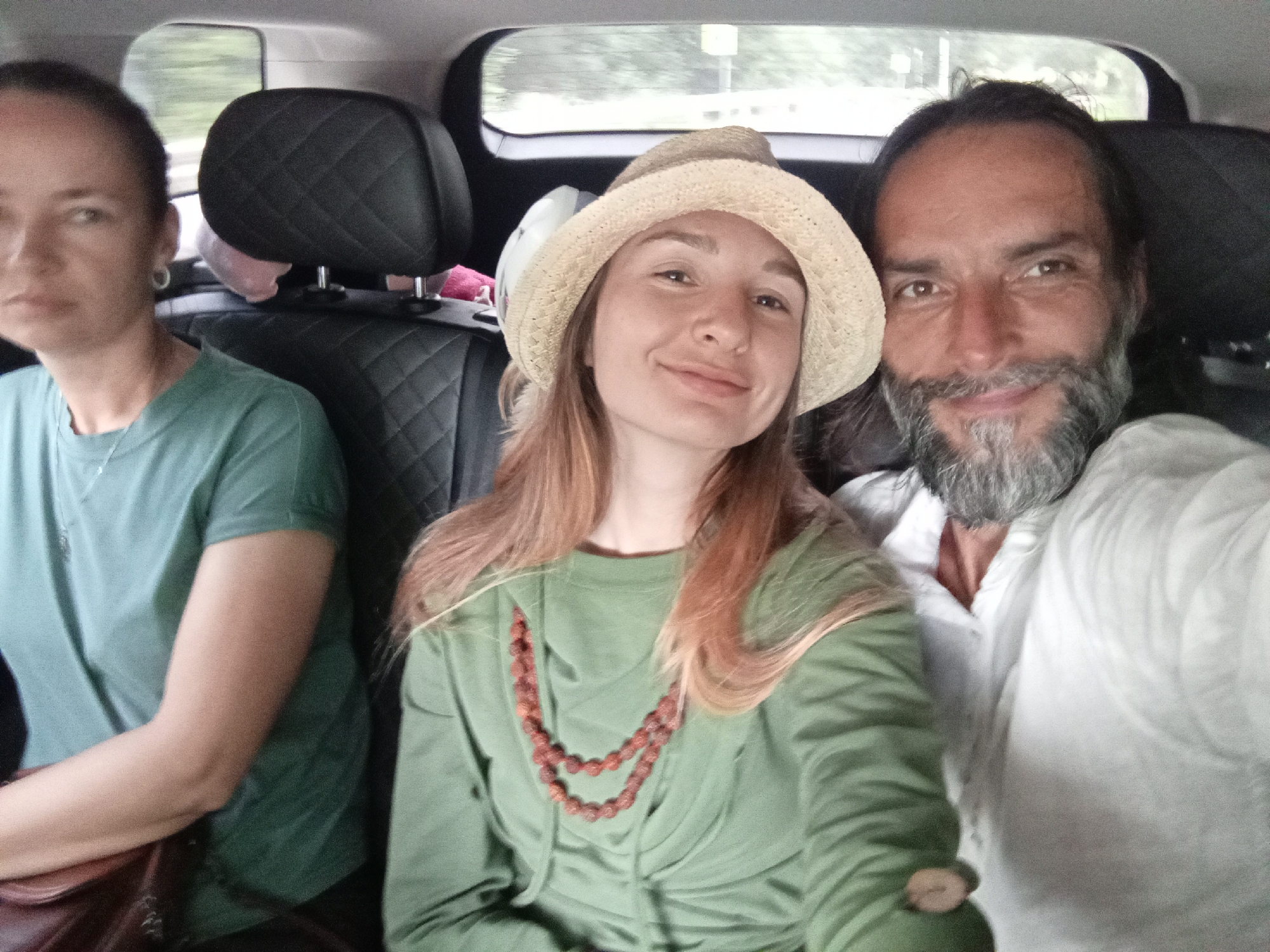 Three people are seated in the back of a vehicle, appearing to be in motion. On the far left is a woman with dark hair pulled back, wearing a teal t-shirt and a silver necklace. To her right, in the center, is a woman wearing a straw hat and a green hooded sweatshirt, with a beaded necklace. On the far right is a man with a beard and long dark hair, wearing a white shirt. The interior of the vehicle features dark upholstery with a quilted pattern on the headrests. The background outside the windows is blurred, suggesting movement. The scene is consistent with being inside a car on a road. The provided location of Голубинское сельское поселение, Ukraine, is noted as the context for this observation.