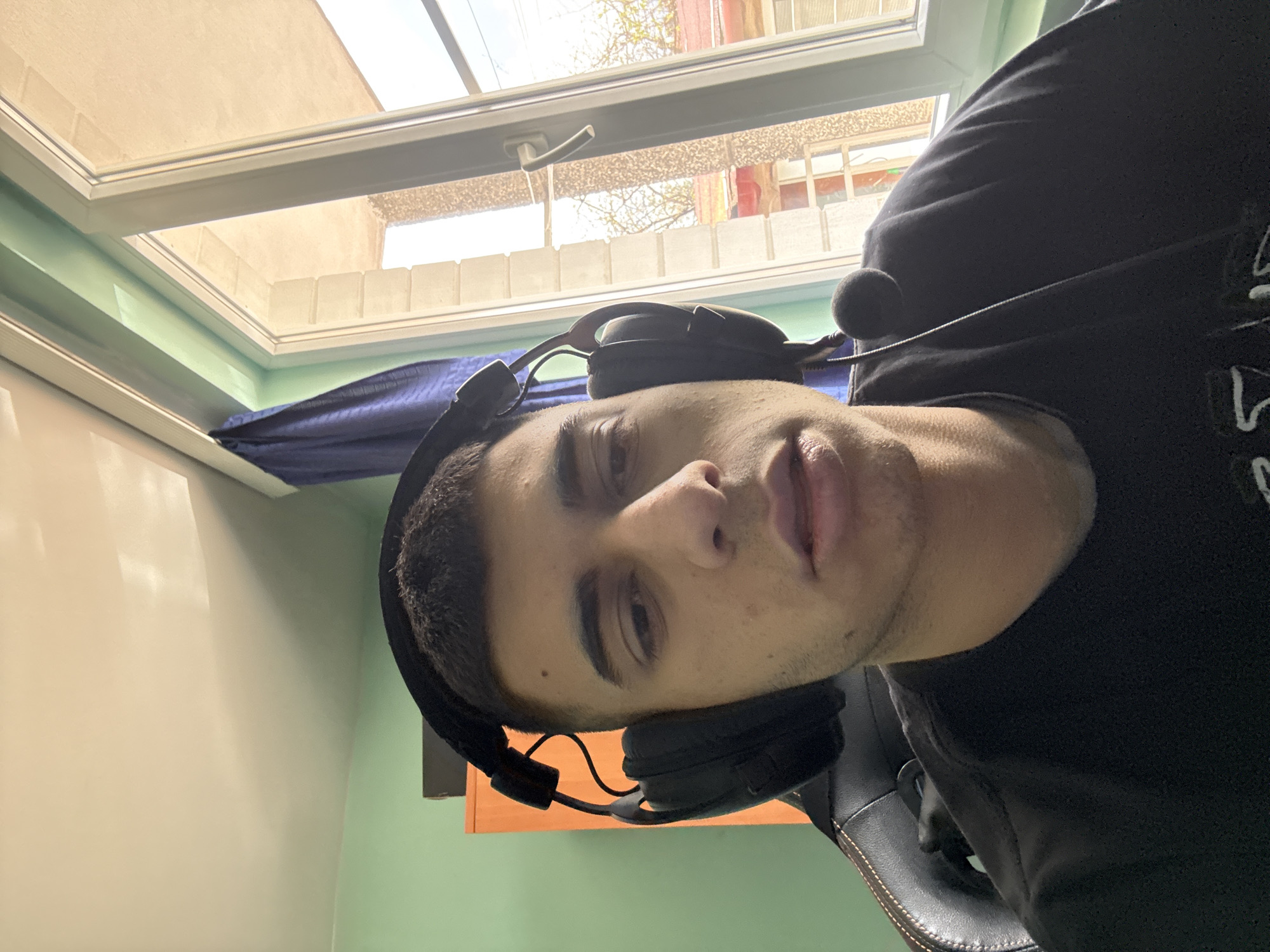 A close-up, rotated headshot of a person wearing black headphones with a microphone. The person is positioned indoors, with a window in the background. The window reveals an exterior scene, potentially in Sofia, Bulgaria, based on architectural details. The surrounding environment consists of a green and white wall, a blue curtain and a leather chair.