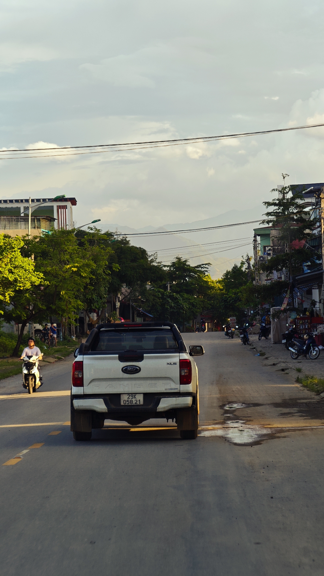 The image captures a street scene in Liên Sơn, Vietnam, under a cloudy, late afternoon or early evening sky with a warm, soft light. A white Ford Ranger XLS pickup truck, license plate "29K 058.21", is centrally positioned, driving away from the viewer on a paved road. The road surface features a faded yellow dashed line on the left and several visible puddles on the right. On the left side of the road, a person on a white scooter is driving towards the viewer. Both sides of the street are lined with dense green trees and multi-story buildings, some featuring green roofs and open verandas. In the distant background, a range of hazy mountains is visible. Multiple black power or communication lines stretch horizontally across the upper mid-ground. Several parked motorcycles are visible along the right side of the street, and small groups of individuals are present near the buildings.