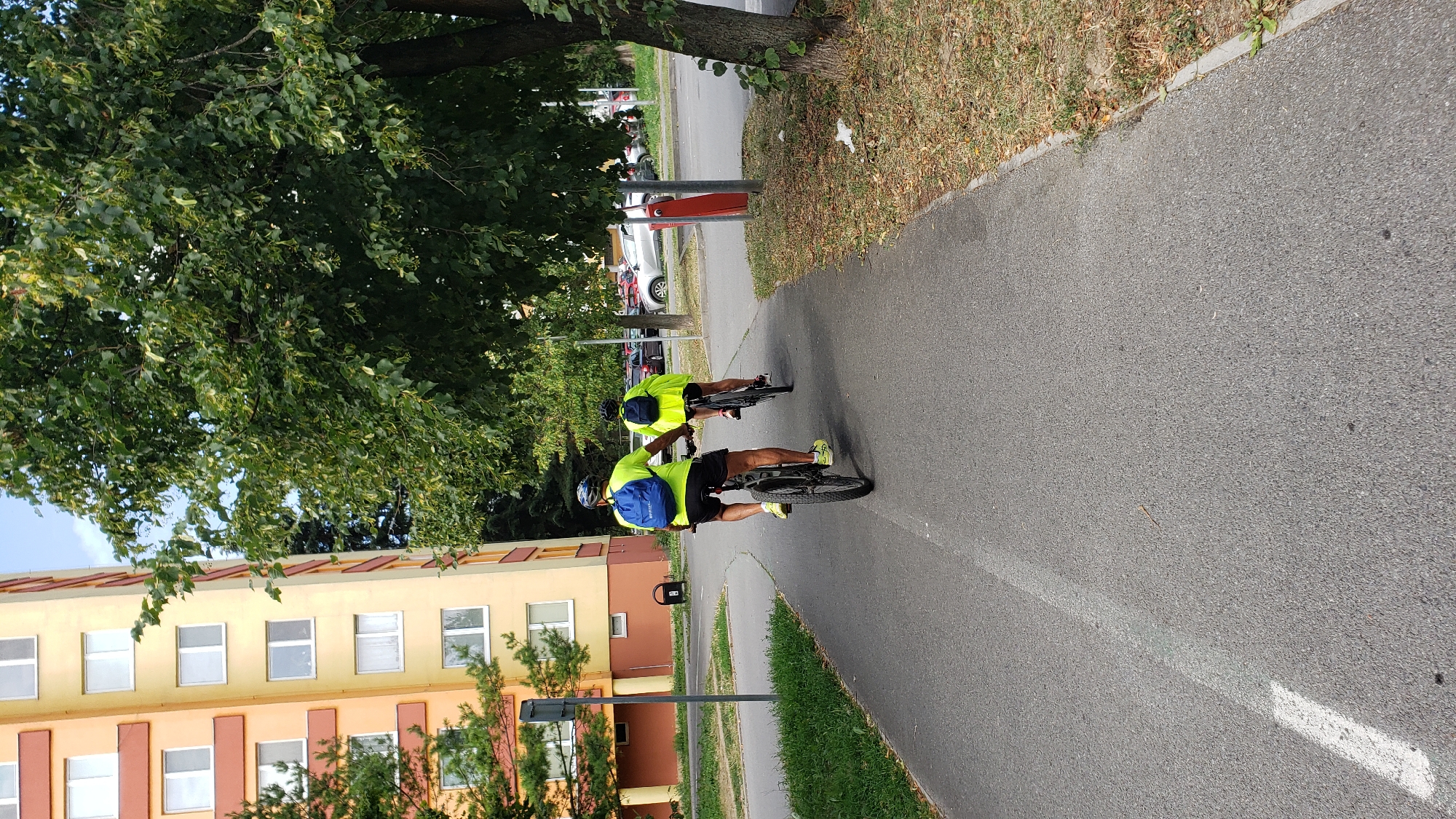 Two individuals are cycling on a paved bike path in Prešov, Slovakia. The person in the foreground, wearing a bright yellow shirt and black shorts, rides a mountain bike. They have a blue backpack on their back and a helmet on their head. The second person, further ahead and slightly to the right, also wears a bright yellow vest and rides a bicycle. This person also has a backpack and is wearing a helmet.
The bike path is asphalt with a white line marking its edge on the left. To the left of the path is a grassy area with some trees. In the background, a tall, multi-story building with orange and yellow siding is visible. To the right of the bike path is a strip of dry grass and fallen leaves, bordered by a concrete curb. Beyond the grass, a street with parked cars is visible. The scene is set under a partly cloudy sky, with lush green trees dominating the right side of the image.
