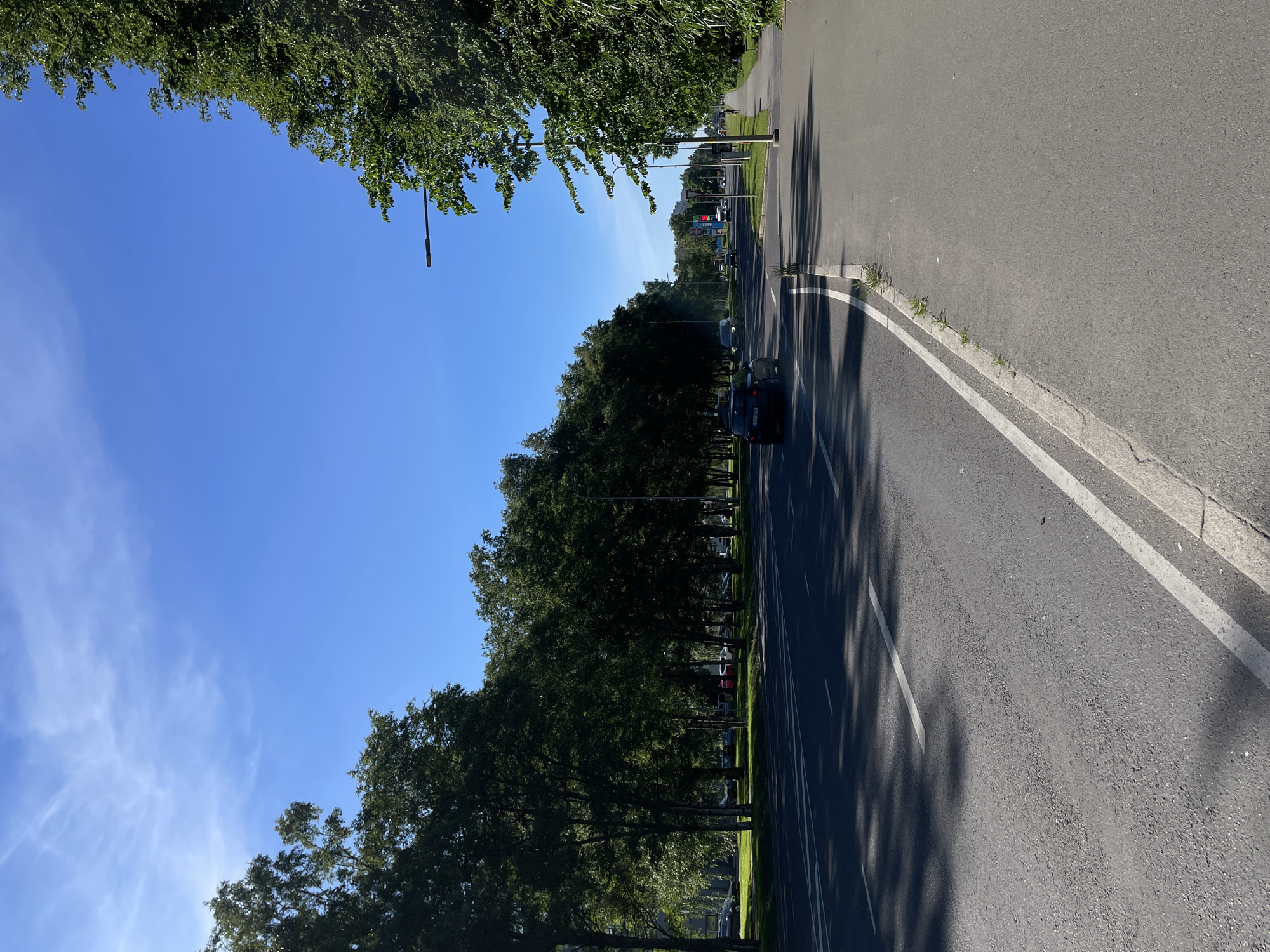 The image depicts a street scene in Tallinn, Estonia, on a bright, sunny day with a clear blue sky and sparse wisps of white clouds. The perspective is from ground level, looking down a paved road lined with lush green trees.
In the foreground, the asphalt road features double white lines, indicating a traffic lane. The sun casts long shadows of the trees onto the road, suggesting it is likely mid-morning or late afternoon. To the left of the road, a dense row of mature trees creates a verdant canopy, with their branches reaching towards the sky. On the right side of the road, there appears to be a sidewalk or paved area, and further to the right, grass and more trees.
Further down the road, several cars are visible, including a dark-colored vehicle in the middle distance. There are also streetlights and signs, one of which displays colorful digital information, possibly advertising or directional. A pedestrian can be seen walking on the sidewalk in the distance.
The overall impression is one of a peaceful, pleasant day in a well-maintained urban or suburban environment, likely a park or a tree-lined avenue. The bright sunlight and green foliage evoke a sense of warmth and tranquility. There is no specific activity or event taking place, but rather a snapshot of daily life. No discernible text is present, beyond what might be on distant signs.