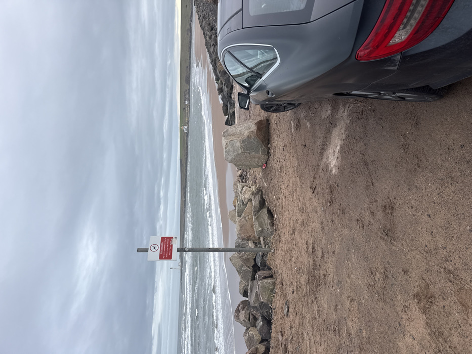 The image presents a coastal scene at Montrose Beach, Montrose, United Kingdom, under an overcast sky. In the immediate foreground on the right, the rear passenger side of a dark-colored two-door coupe vehicle is partially visible, parked on a reddish-brown dirt and sandy surface exhibiting tire tracks. To the left of the vehicle, a formation of large, irregular rocks acts as a natural barrier, with a notably large, light-colored boulder positioned centrally among them. A grey metal pole rises from this rocky area, supporting a rectangular white sign with a red border. The sign displays a red 'no dogs' pictogram and the prominent text "DOG CONTROL," followed by smaller, partially legible regulatory text.
Beyond the rocks and sign, the North Sea is visible with breaking waves approaching a sandy beach. On the distant horizon to the left, a tall, white lighthouse (North Esk Lighthouse) stands. Further along the coast to the right, a smaller white lighthouse, accompanied by white buildings (Montrose Lighthouse), marks the entrance to a body of water, likely the Montrose Basin or River South Esk. The distant coastline consists of low-lying, green-brown hills. The sky is uniformly grey with heavy cloud cover. No individuals are present in the scene.