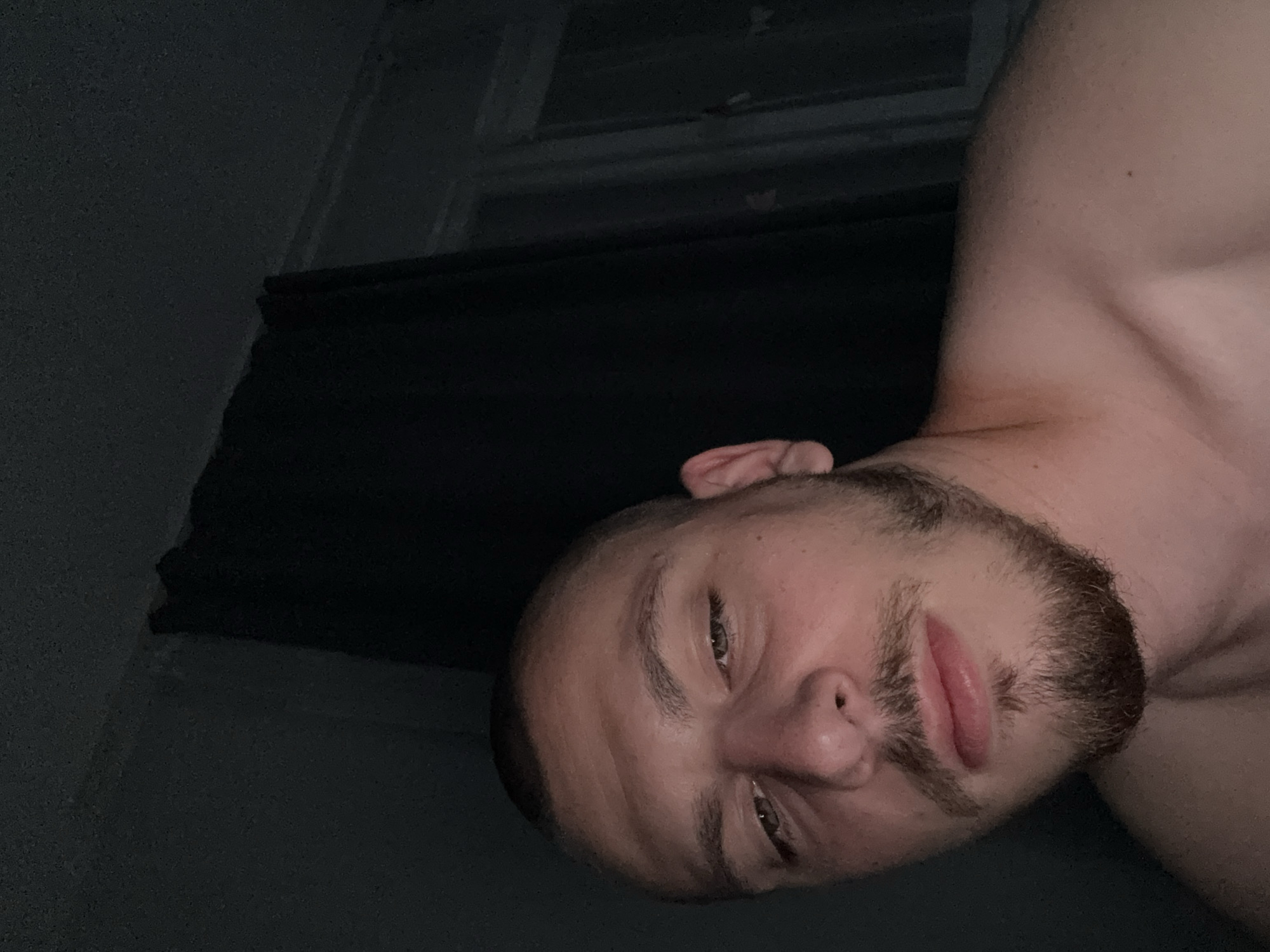 The image is a close-up, low-angle selfie taken indoors, likely in a bedroom, given the presence of a window with blinds and dark curtains. The lighting is dim, suggesting it was taken at night or in a low-light environment. The subject is a man with a shaved head and a beard. He is looking slightly down and to his left, with his eyes partially closed. His skin appears flushed. His bare shoulder and upper chest are visible. The background consists of dark curtains and a window with diffused light. There is no information in the image to suggest the location is Stara Zagora, Bulgaria.