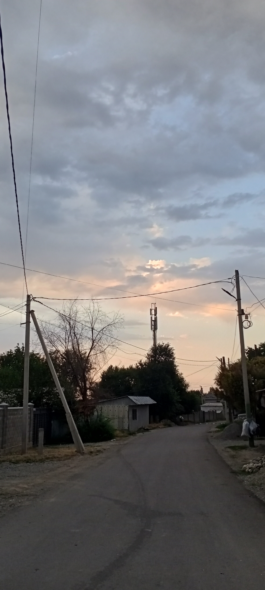 The image depicts a street scene in the city of Sokuluk, Kyrgyzstan, during what appears to be dusk. A paved road curves gently to the right, with tire marks visible on its surface. On the left side of the road, a concrete fence with vertical posts encloses a property, and a leaning utility pole supports overhead wires. Further down the road on the left, a small, dilapidated shed with a corrugated metal roof stands near some vegetation. A tall cell tower is visible in the mid-ground, amidst a cluster of trees. On the right side of the road, another utility pole is present, and piles of gravel and rocks are visible. The sky is filled with cloudy formations, with hints of pink and orange suggesting the setting sun breaking through. There are no people visible in the image.