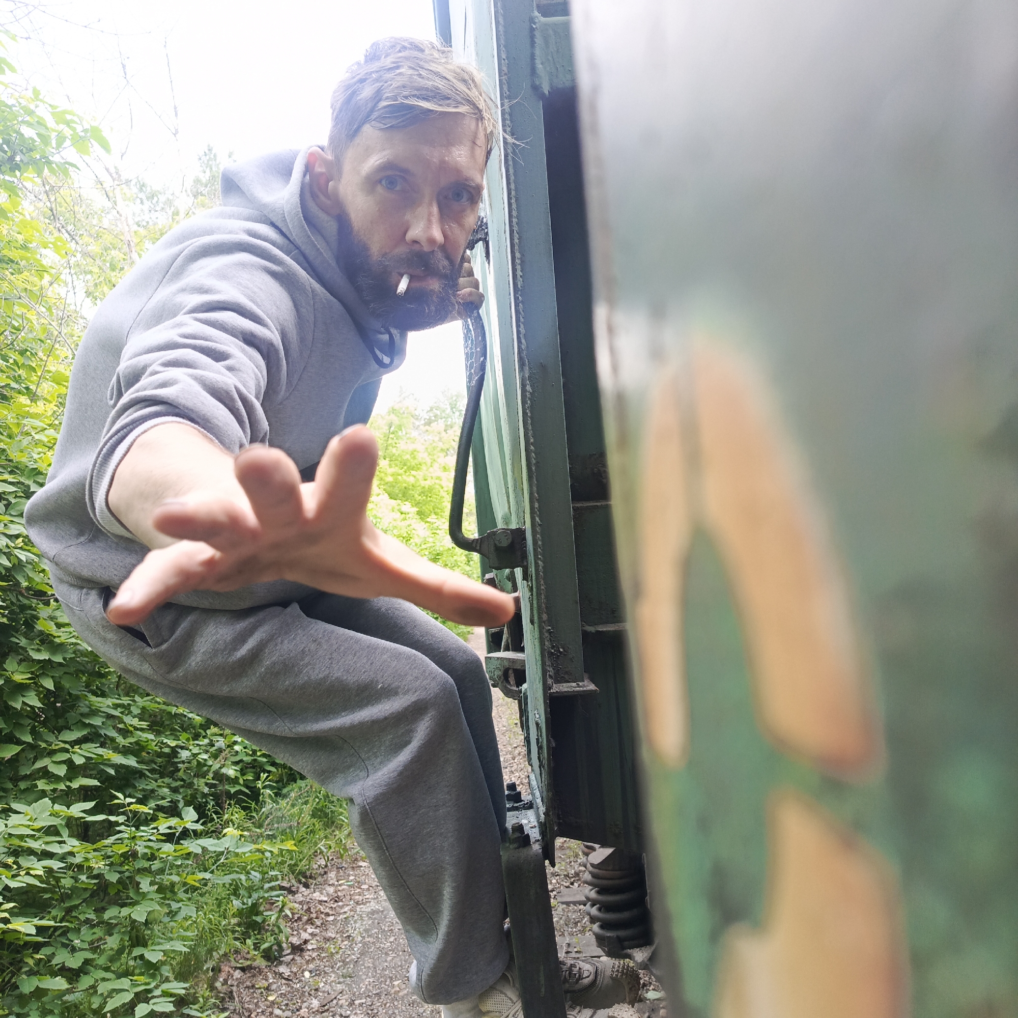 In Chaikovsky, Russia, a man with a beard and a cigarette in his mouth is pictured from a low-angle perspective, reaching out his left hand towards the camera. He is wearing a grey hooded sweatshirt and grey sweatpants. He is seated on the edge of a green, weathered train car. The train car has visible bolts and a metal handle. Green foliage and trees are visible in the background, suggesting an outdoor, possibly rural or semi-rural location. The ground appears to be a dirt path with some gravel. The overall scene captures a candid moment with the man in motion, his hand extended in a gesture that could be interpreted as an invitation or a warning.