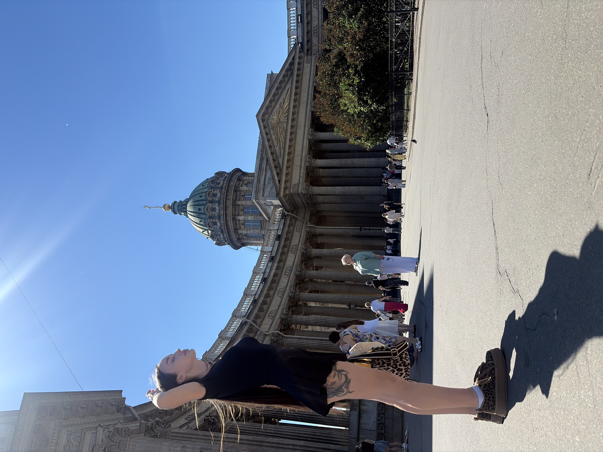 The image captures a sunny day in Saint Petersburg, Russia, likely in front of Kazan Cathedral. In the foreground, a young woman with long dark hair, wearing a black dress and leopard-print platform sneakers, stands with her hands behind her head, looking to her left. She has extensive tattoos visible on her left leg. To her right, a group of people are walking on a paved plaza, with the grand facade of Kazan Cathedral forming the background. The cathedral features a prominent dome and a colonnade. The sky is clear and blue, with sunlight casting distinct shadows on the ground. Several people are seen in various distances, some appearing to be tourists or locals enjoying the public space. A small bird is visible on the ground in the middle distance.