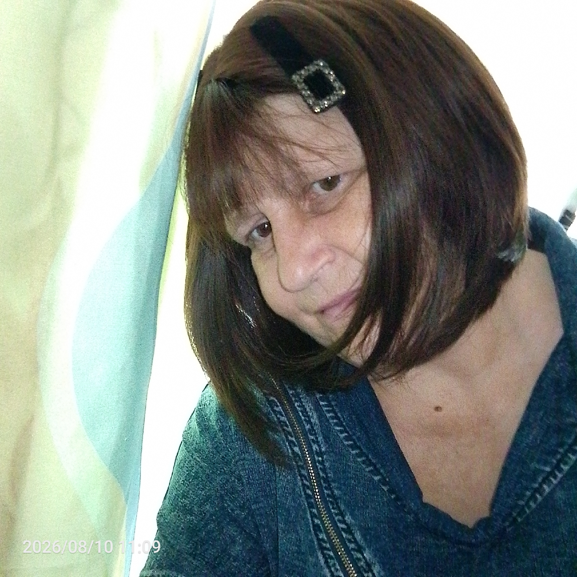 The image is a close-up, medium-shot, self-portrait of a middle-aged woman with brown hair and bangs. She is wearing a dark blue denim jacket with a zipper closure. A black headband with a square, jeweled accent is visible in her hair. Her left eye is looking directly at the camera. The background consists of light-colored fabric, possibly a curtain or drapery, with a blue-green overlay. A timestamp "2026/08/10 11:09" is visible in the lower left corner of the image. The setting is indoors, with soft lighting.