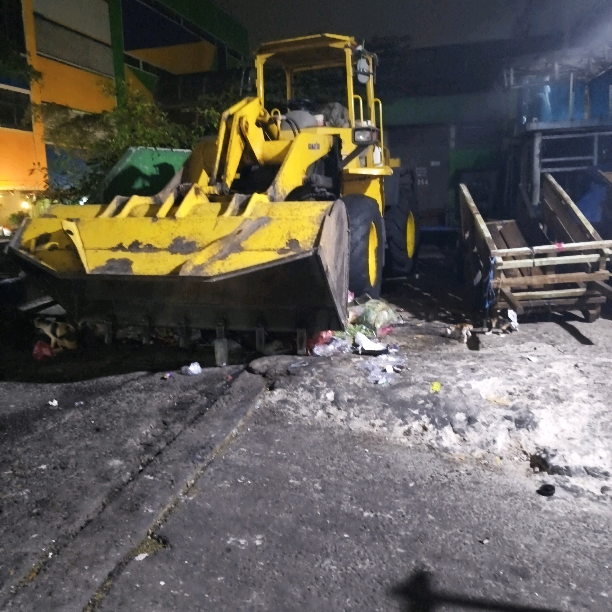 A yellow front-end loader is parked on an asphalt surface, possibly in a waste management or industrial area in South Jakarta, Indonesia. The loader has its bucket lowered, resting on the ground. Several piles of trash and debris are visible around the loader, including plastic bags and other refuse. In the background, there are structures that appear to be buildings or part of a facility, with some green and orange elements. The lighting suggests it is nighttime or a poorly lit area. The scene appears to be a work in progress or a site where materials are being managed.