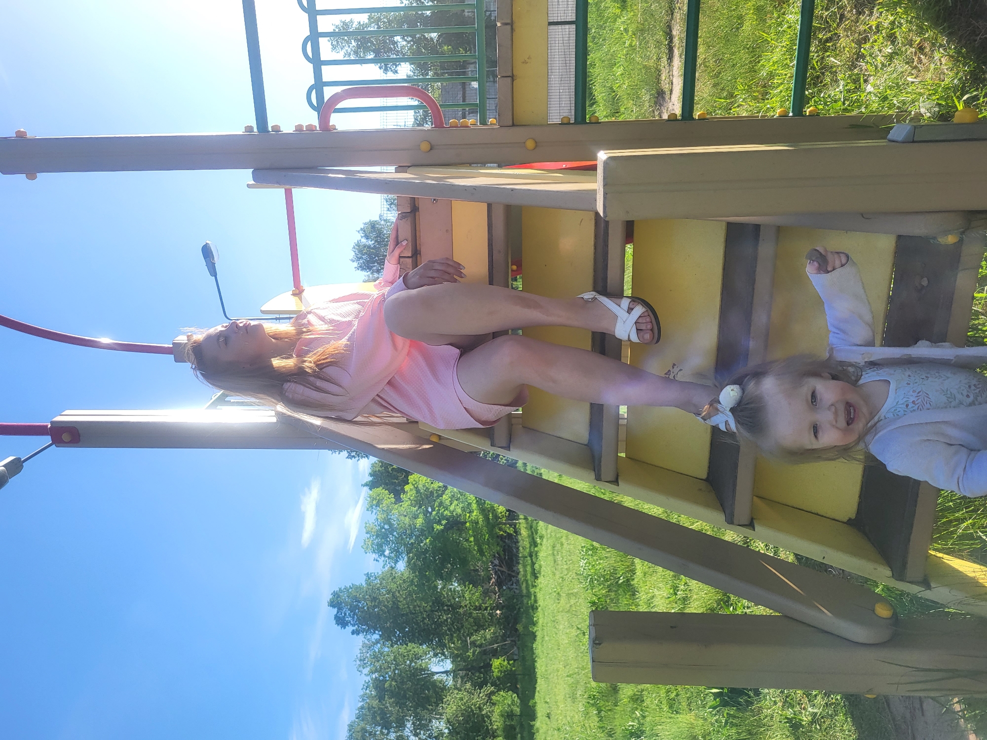 A woman and a young child are on a playground structure. The woman is lying on a section of the structure. She is wearing a pink shirt and white sandals. The child is standing near the woman, looking at the camera. The child has a cast on their left arm. The playground is located in a grassy area with trees in the background. The sky is blue and sunny. It is unknown if this photograph was taken in Chaikovsky, Russia.