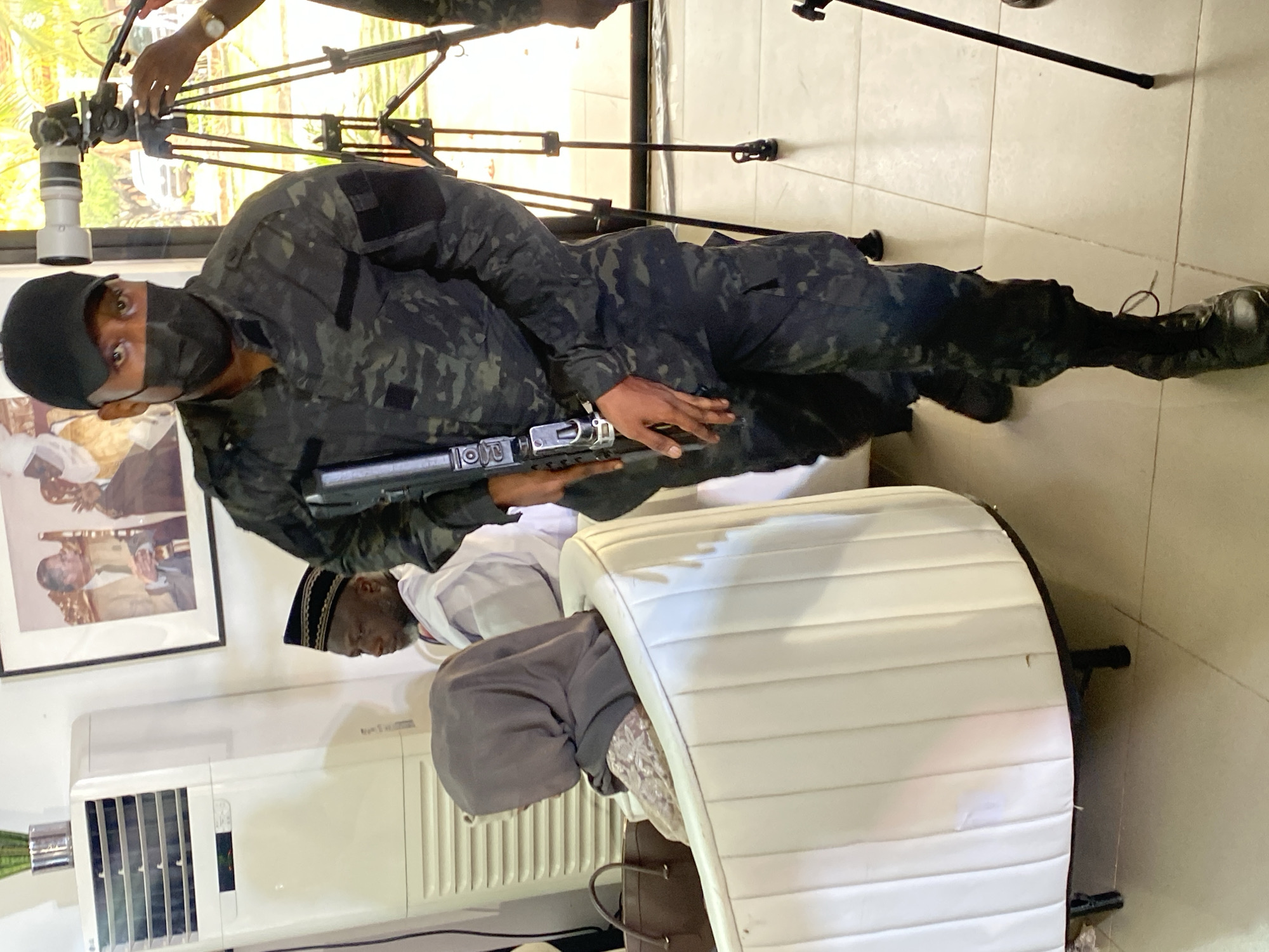 A man wearing a black camouflage uniform, a black mask, and a black cap is lying on the floor. He is holding a silver and black rifle. Behind him are two men sitting on a white tufted sofa. One of the men is wearing a white shirt and a black and gold cap. The other man is wearing a gray hoodie. Behind them is an air conditioner, a framed picture, and a white sofa. In the background is a camera on a tripod. The photograph was taken in Kwarau, Nigeria.