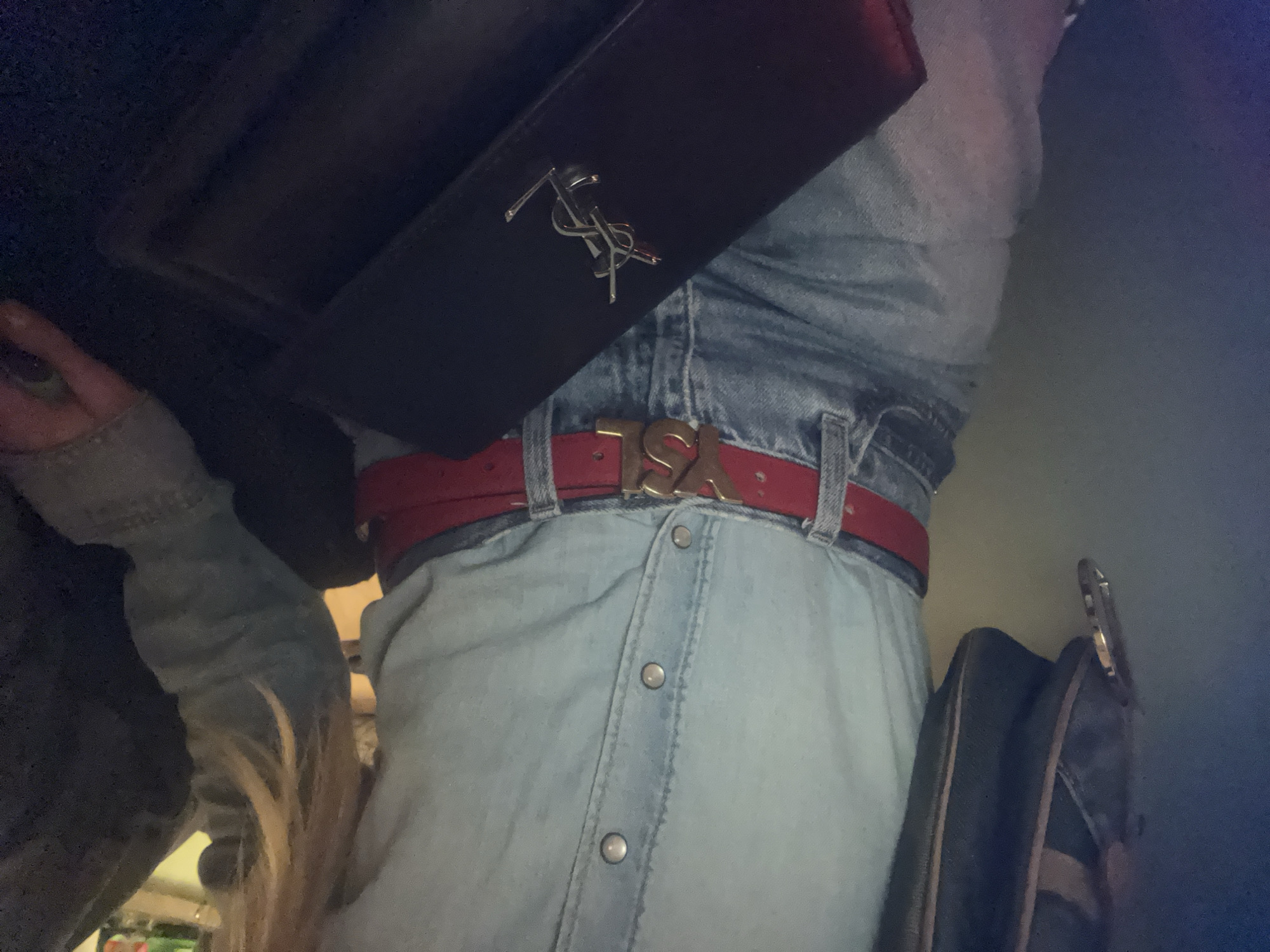 The image shows a person wearing a light blue denim dress with a red belt. The belt has a gold-colored buckle featuring stylized letters. A black rectangular clutch with a silver "YSL" logo is positioned near the top of the dress. The person is also wearing a denim jacket. There is a dark-colored bag with gold hardware on the right side of the frame. The background is unclear, suggesting an indoor setting.