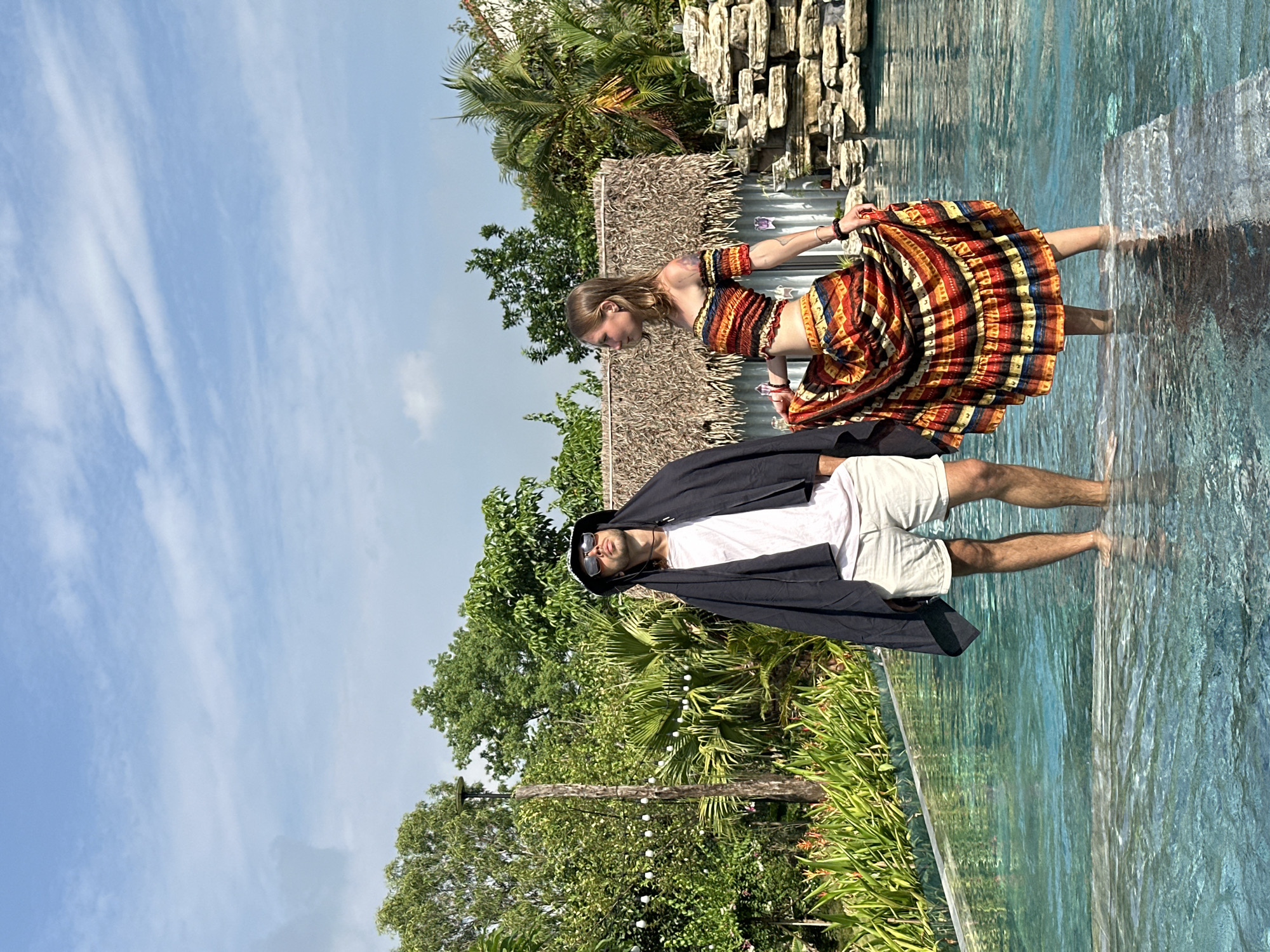 In Nam Hoa Lư Ward, Vietnam, two individuals stand waist-deep in a pristine infinity pool, their feet submerged in the clear, turquoise water. Lush green foliage and tall trees surround the pool, with a backdrop of a thatched-roof structure and a rocky waterfall feature. Above, a bright blue sky is dotted with wispy white clouds.
On the left, a man wearing sunglasses and a dark hat stands with a black cloth draped over his head and shoulders. He is dressed in a light pink t-shirt and light-colored shorts.
To his right, a woman in a vibrant, off-the-shoulder crop top and a matching full skirt with a colorful, patterned design poses with her hand lifting the skirt. Her hair is blonde, and she is adorned with bracelets on both wrists. The woman has a tattoo on her left arm.