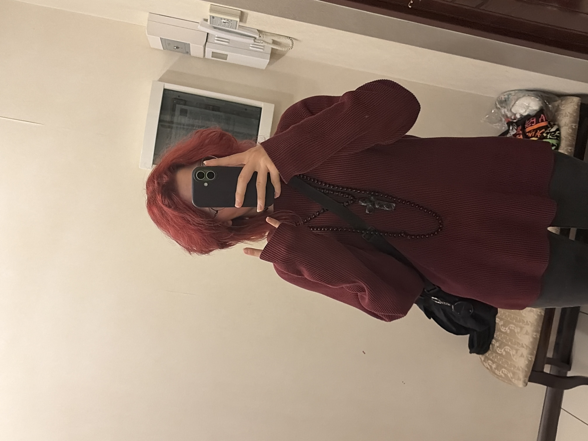 A person with bright red hair is taking a mirror selfie in a room. They are wearing a dark red ribbed sweater, dark pants, and a beaded necklace with a cross pendant. A black bag is slung over their left shoulder and rests on a stool next to them. The background shows a light-colored wall with a framed picture or mirror, a wall-mounted telephone, and a doorway. There are no identifying features to indicate the location is in Chelyabinsk, Russia.