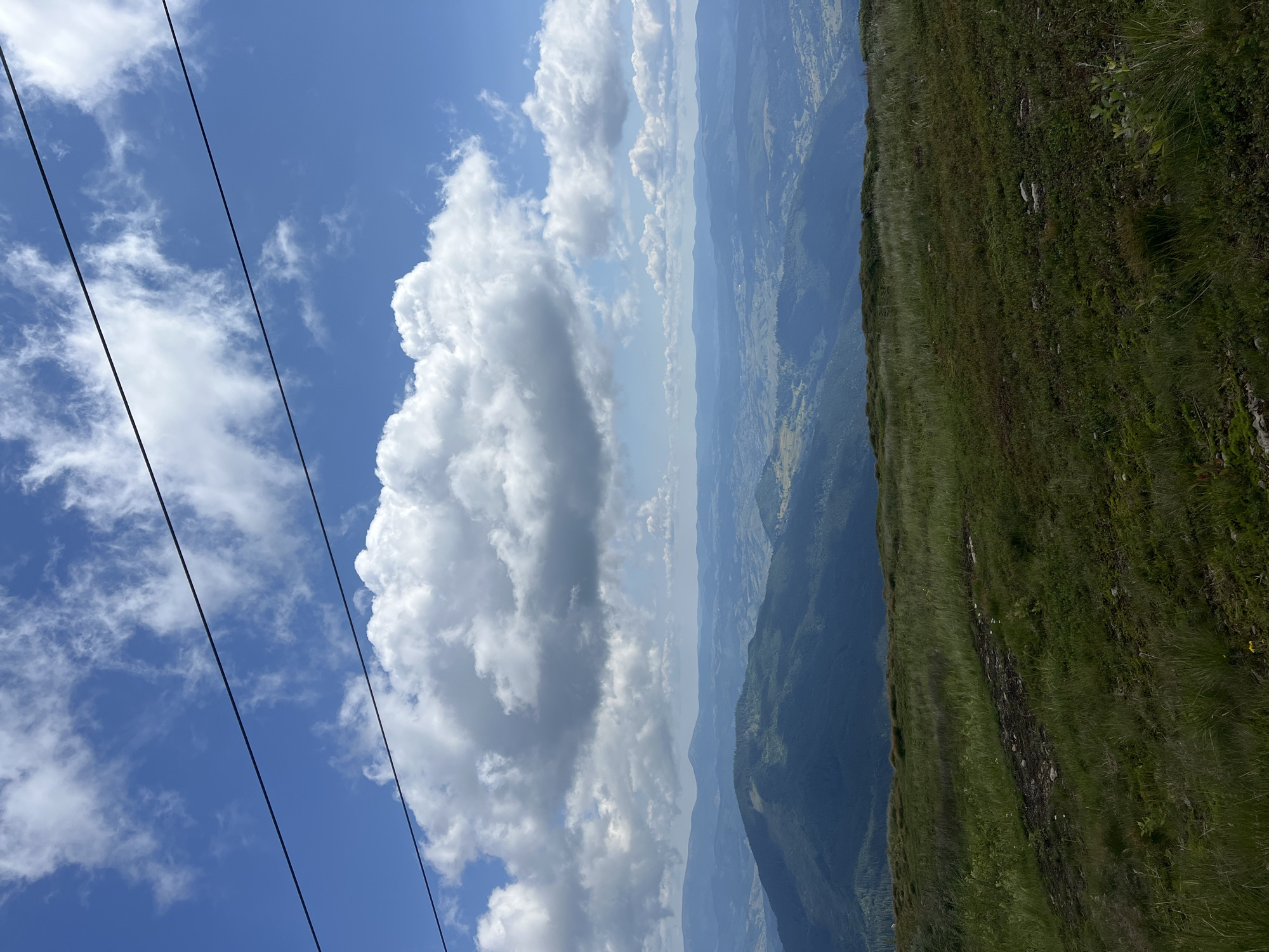 The image depicts a panoramic view of mountainous terrain under a bright blue sky filled with fluffy white clouds. In the foreground, a grassy slope leads down towards a vast expanse of layered, dark blue mountain ranges stretching to the horizon. The mountains are covered in dense vegetation, with some areas appearing as shadows. The sky is dominant in the upper portion of the image, with three dark, parallel lines running diagonally across it, suggestive of power lines or a ski lift cable. The scene appears to be a natural, undeveloped landscape. There are no people or animals visible. The lighting suggests it is daytime, likely late morning or early afternoon.
