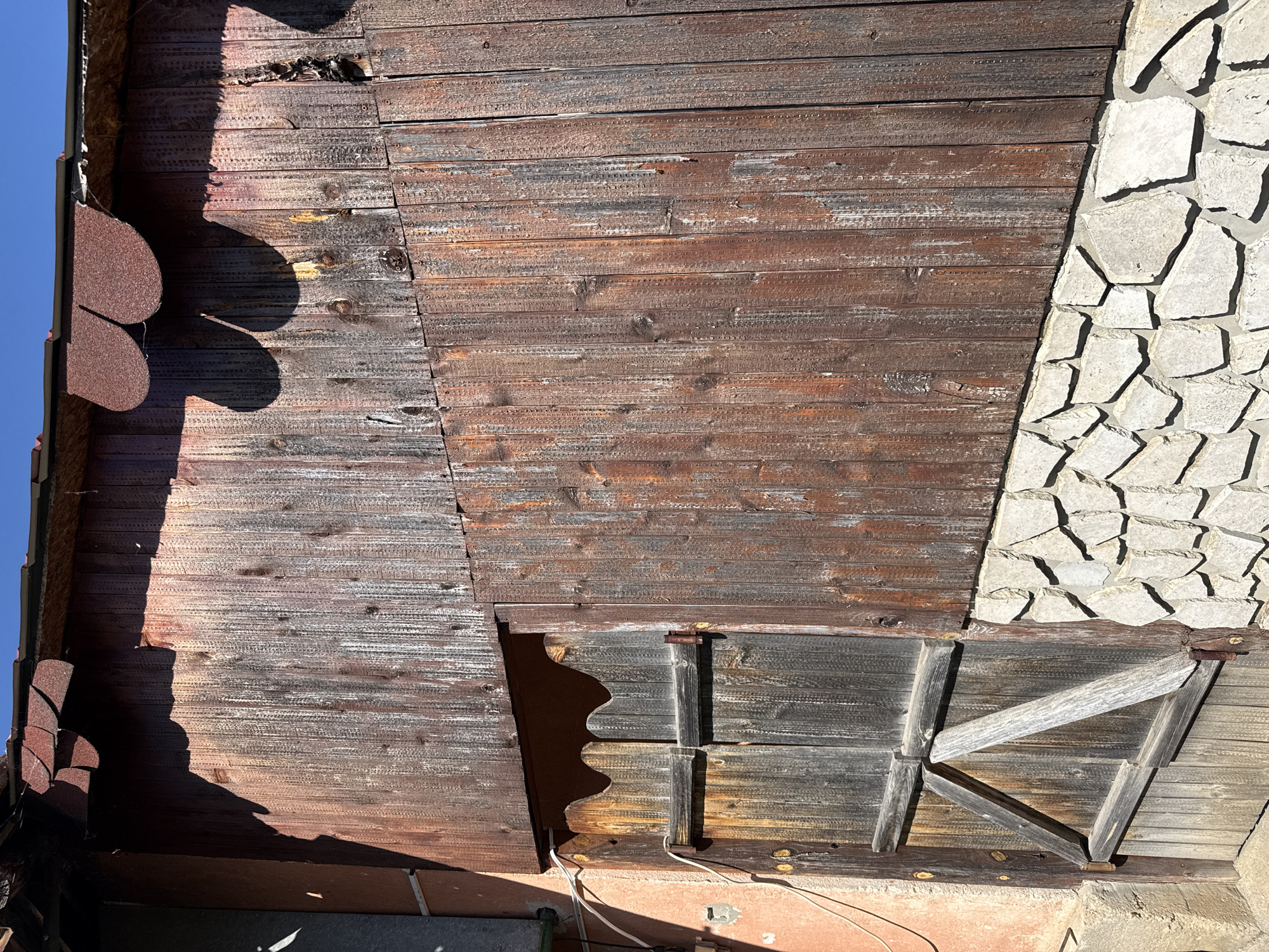 The image shows the exterior of a building in Valchedram, Bulgaria. The wall is composed of weathered, horizontal wooden planks, except for a section on the right side which is covered in light-colored, irregularly shaped stones. There is a small, wooden shuttered window near the bottom. The roofline is partially visible with decorative red tiles along the edge. A clear blue sky can be seen in the upper left corner.