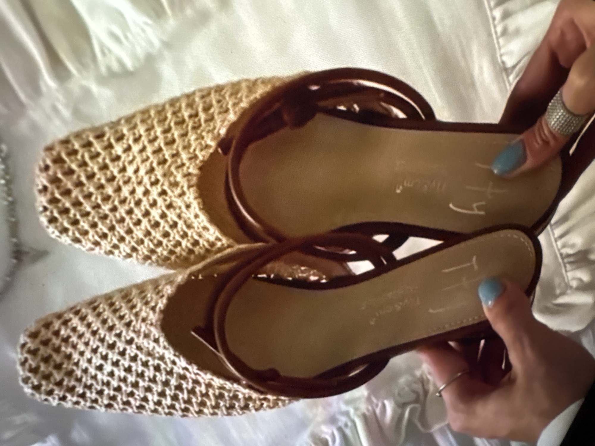 The image shows a pair of flat sandals held up in someone's hands. The sandals have a woven texture on the upper part, and a brown leather sole with the brand name "TOMS" printed on it, alongside some handwritten initials. The hands holding the sandals have manicured nails, one with a silver ring and the other with a ring made of small stones. The background appears to be a white fabric with a subtle texture. The location is not identifiable from the image.
