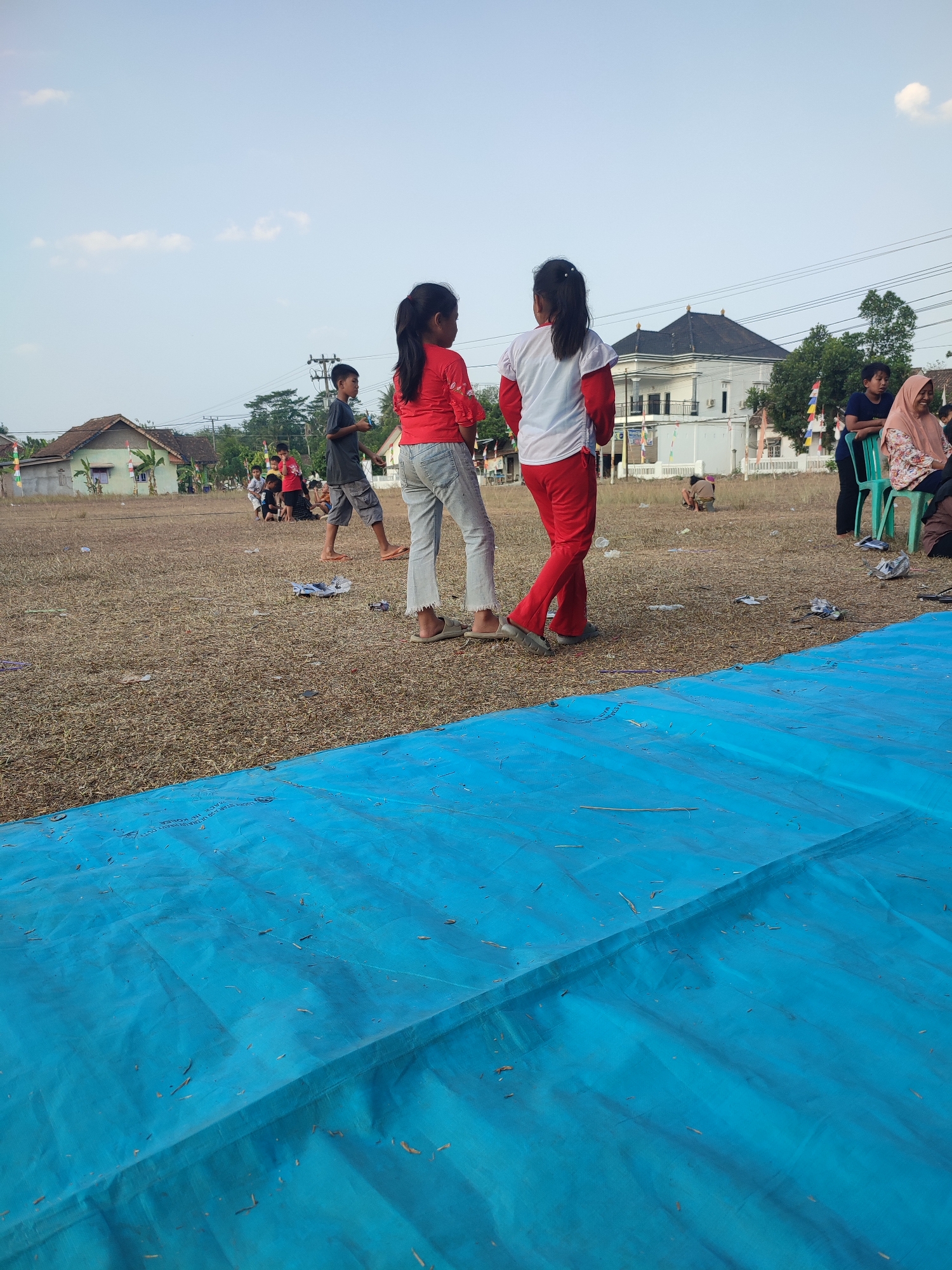 The image captures an outdoor scene in a field at Nusa Jaya, Indonesia, characterized by dry, yellowish grass and scattered debris. In the foreground, a large, bright blue tarp dominates the lower portion of the frame. Behind the tarp, two young girls are the primary subjects. The girl on the left wears a red long-sleeved shirt and light-colored pants. The girl on the right is dressed in a white long-sleeved shirt and red pants. Both are facing away from the camera, towards the middle ground and background.
Further back, a group of people are visible, including several children and some adults. A boy in a dark t-shirt and shorts stands in the middle ground. To his right, an adult in a light pink headscarf and patterned shirt sits on a green chair. A white building with a dark roof and windows is situated in the background, along with other houses and trees. Power lines are also present, stretching across the sky. The sky is light blue with a few scattered clouds. The overall impression is of a community gathering or event taking place in a somewhat unkempt open area.