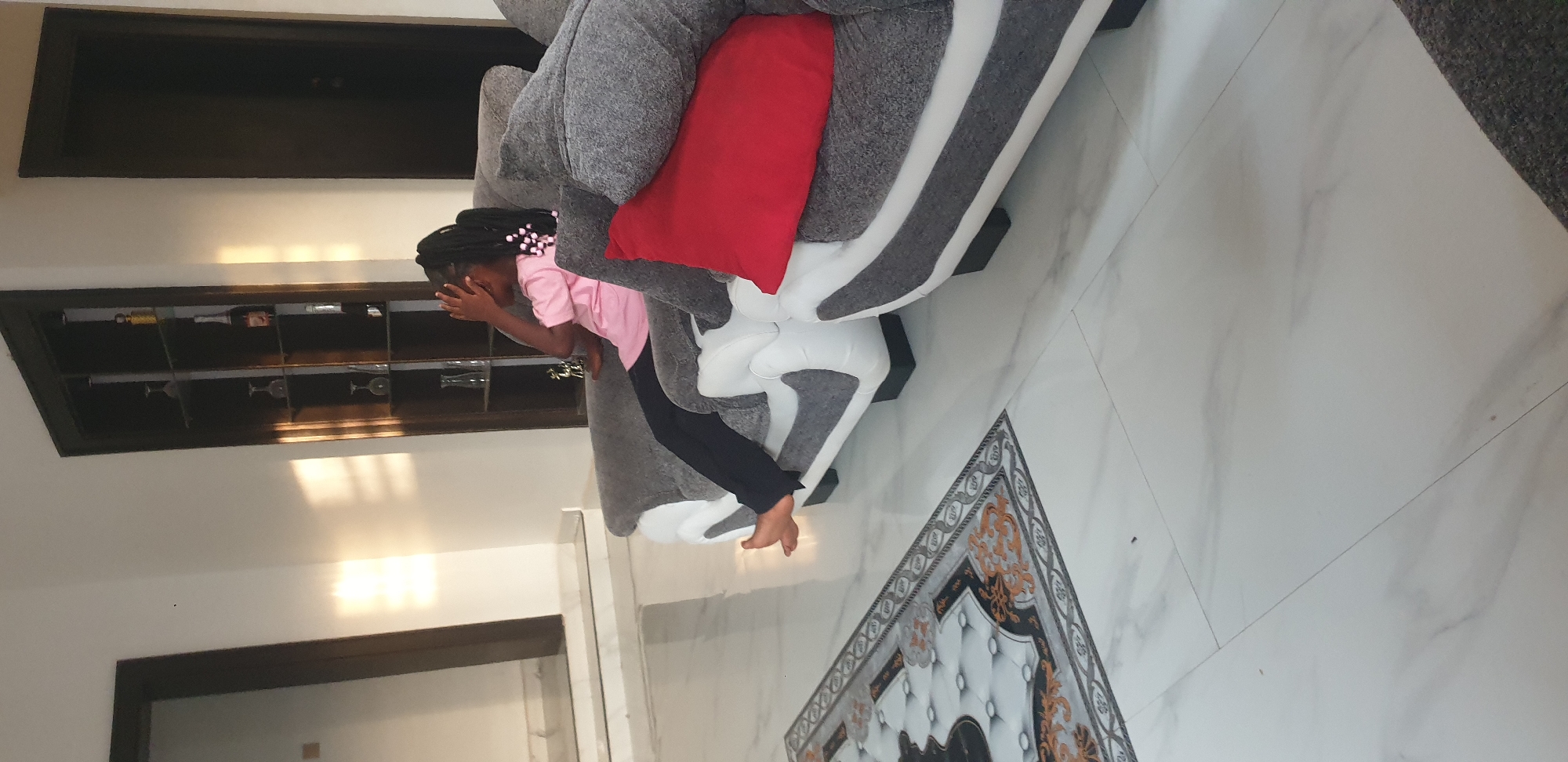 The scene depicts an indoor living area, reportedly in Barden Yana, Nigeria. A young individual with braided hair adorned with white and pink beads is seated on a modern sofa. The individual wears a pink short-sleeved top and dark pants, is barefoot, and uses their right hand to cover their face, with the left hand resting on the grey and white upholstered sofa. A red throw pillow rests on the sofa to the right. The floor is covered with polished white marble or large format tiles featuring subtle grey veining. A decorative rectangular mat with a black, white, and gold pattern is partially visible on the left side of the floor. In the background, a tall, dark wooden display cabinet with glass shelves contains various items including glassware (wine glasses, bottles, vases) and a small golden figurine. Flanking this cabinet are two dark brown doors. Sunlight casts distinct patches of light and shadow on the white walls, illuminating the space.