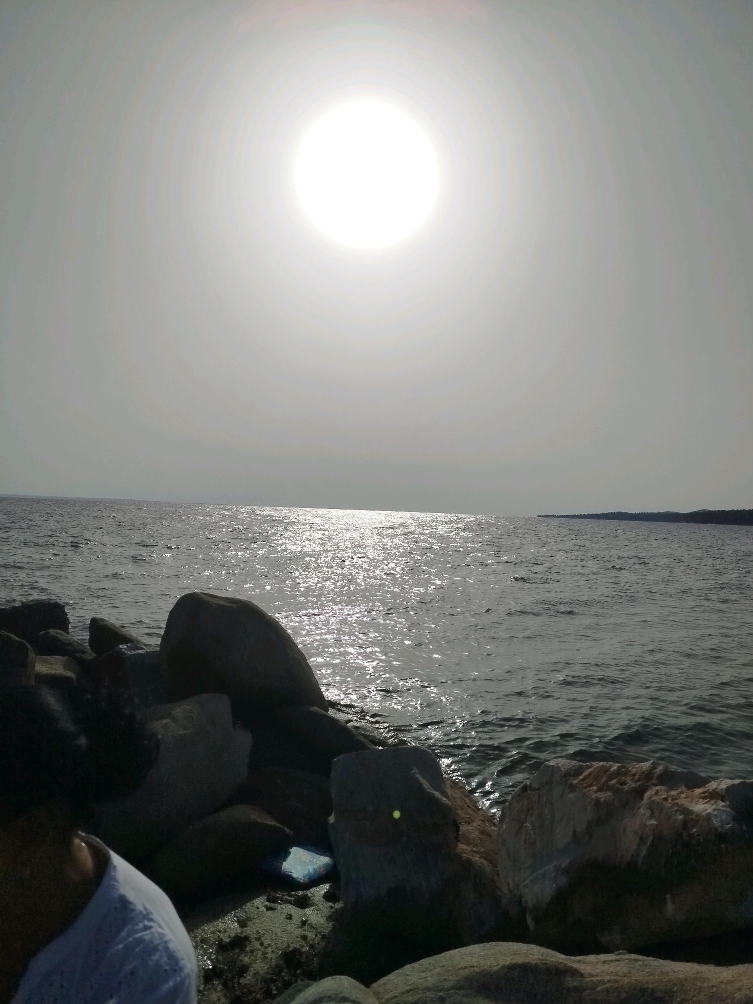 The image shows a bright, overcast sky with the sun visible as a large, diffused circle in the upper portion. Below the sky is the sea, with sunlight reflecting off its surface, creating a shimmering effect. In the foreground, there are large rocks, some of which are dark and silhouetted. A person's head and shoulder are visible on the left side of the frame, wearing a white, textured top. The scene appears to be a coastal area. The location is the Sithonia Municipal Unit, Greece.