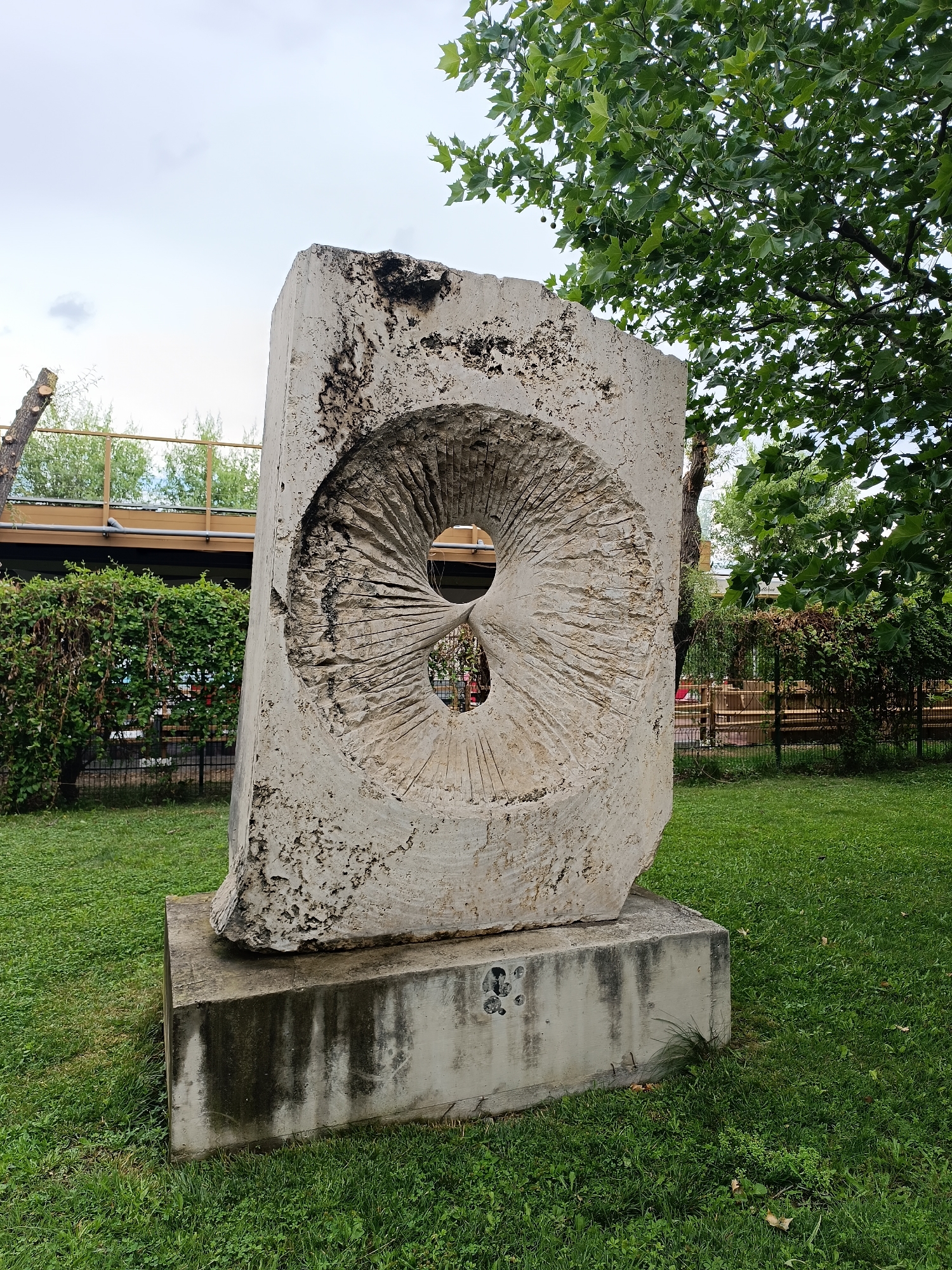 The image shows a large, rough-hewn stone sculpture with a circular opening carved into its center, set on a concrete base in a grassy park. The sculpture is weathered and textured, with dark marks and chips on its surface. The opening in the sculpture is also textured with radiating lines, creating a sense of depth. In the background, there is a lush green hedge, a metal fence, and trees with broad leaves. Above the hedge, a wooden structure with railings is visible, suggesting a raised walkway or building. The sky is overcast. This sculpture is located in the city of Târgu Jiu, Romania, and is part of the "Ensemble of Constantin Brâncuși" sculptures, specifically identified as "The Portal of the Kiss."