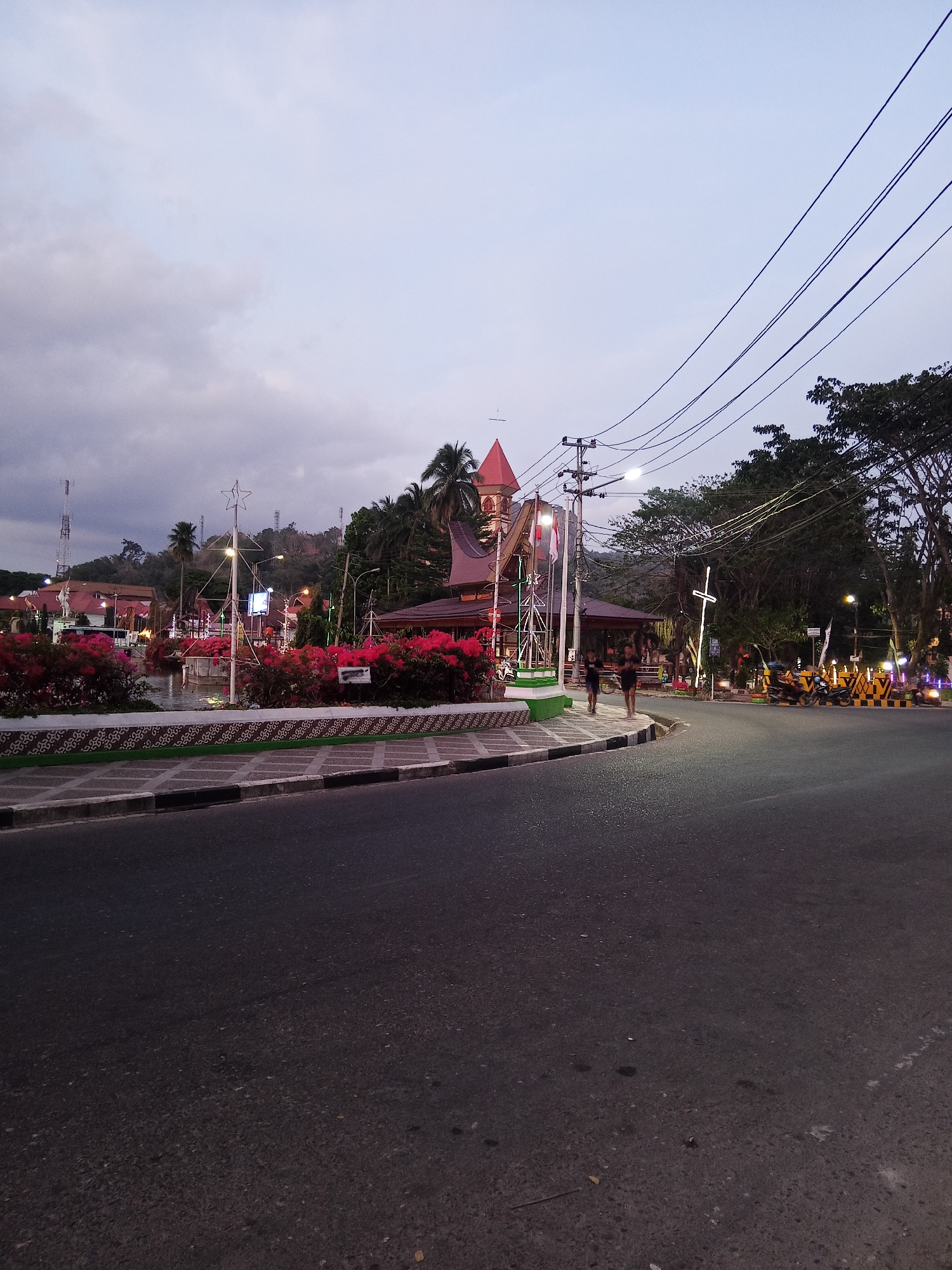 The image depicts a street scene in Makale, Indonesia, at dusk. In the foreground, a paved road curves to the right. On the left side of the road, there is a sidewalk with a decorative border, followed by a landscaped area with vibrant pink flowers. Beyond the flowers, a body of water is visible, with a Christmas tree-like structure adorned with lights in the center. Several buildings are present in the background, including one with a red-roofed tower that resembles a church. Overhead, power lines stretch across the sky. Two individuals are walking on the sidewalk. Further down the street, several motorcycles are parked, and decorative lights illuminate the area, suggesting a festive atmosphere. The sky is overcast with a soft, diffused light.