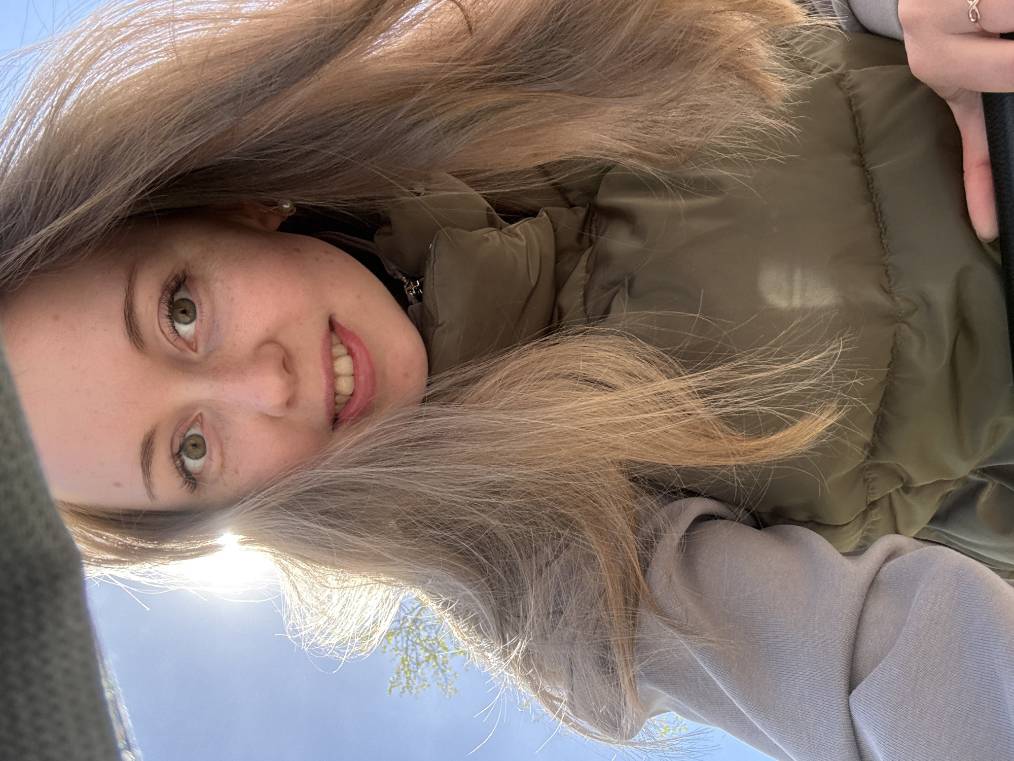 A young woman is looking down at the camera. She has blond hair and green eyes. She is wearing a green puffer jacket. There are trees in the background. The location is unknown.