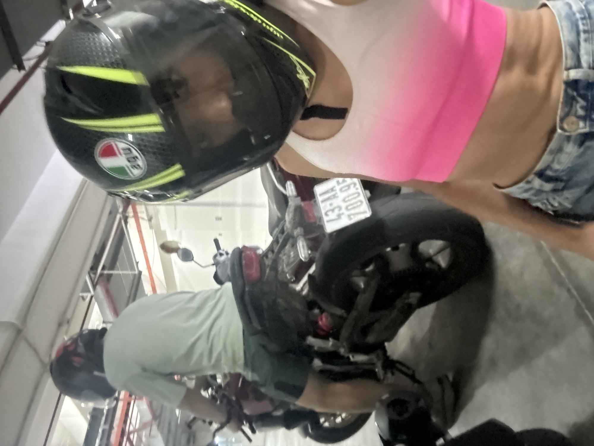 The image captures a tilted, high-angle view inside what appears to be an indoor parking garage with concrete flooring and overhead pipes.
In the immediate foreground, a person is partially visible, wearing a black motorcycle helmet with neon yellow stripes and an AGV brand logo. Their face is obscured by the helmet's dark visor. They are dressed in a pink sleeveless crop top and blue denim overall-style shorts, with a bare midriff.
Behind and below this person, another individual is seen bent over a motorcycle. This person wears a light green t-shirt and dark shorts or trousers, and a dark red/black helmet is on their head. They seem to be interacting with the motorcycle, possibly mounting or dismounting. The motorcycle they are with has a license plate clearly displaying "43-AA 700.95", which is consistent with the Đà Nẵng, Vietnam location context. Part of a second motorcycle is visible beneath the foreground person.
The overall activity suggests people are preparing to ride or have just finished riding motorcycles. The setting is indoors, with artificial lighting, so the time of day and weather are not discernible. No specific emotions are visible.