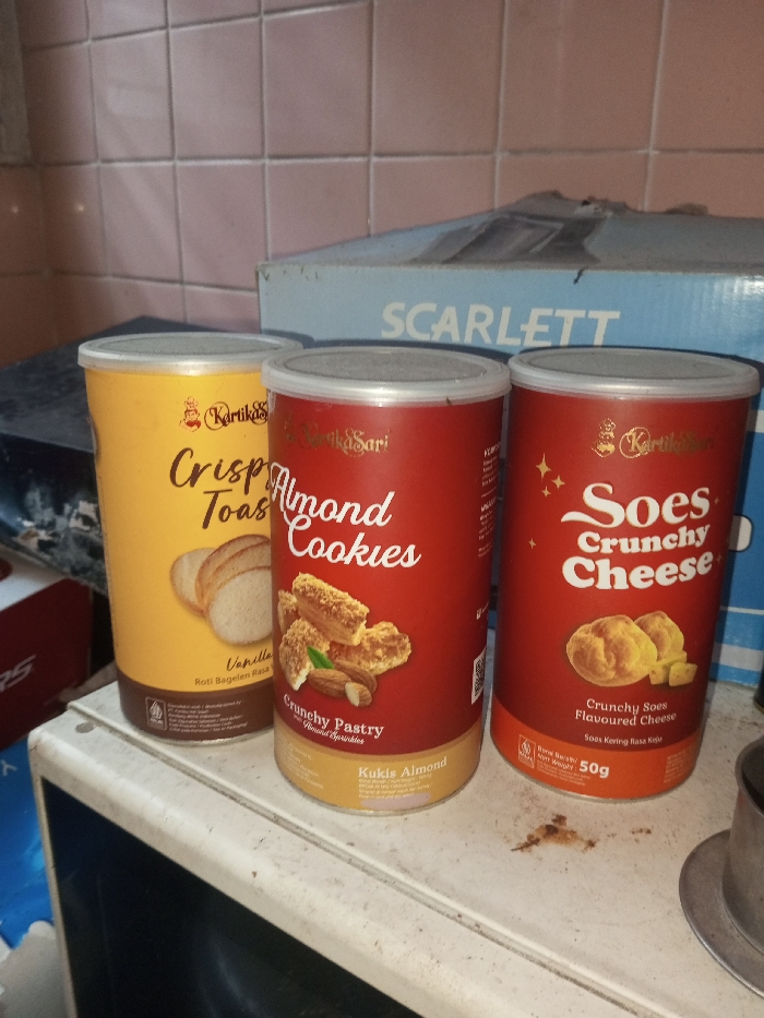 The image showcases three cans of Kartika Sari cookies. From left to right, they are "Crispy Toast" with a vanilla flavor, "Almond Cookies," labeled as "Crunchy Pastry," and "Soes Crunchy Cheese," described as "Crunchy Soes Flavoured Cheese." The Soes Crunchy Cheese can indicates a net weight of 50g. A Scarlett box is visible in the background, and the cans are placed on a surface with some visible dirt.