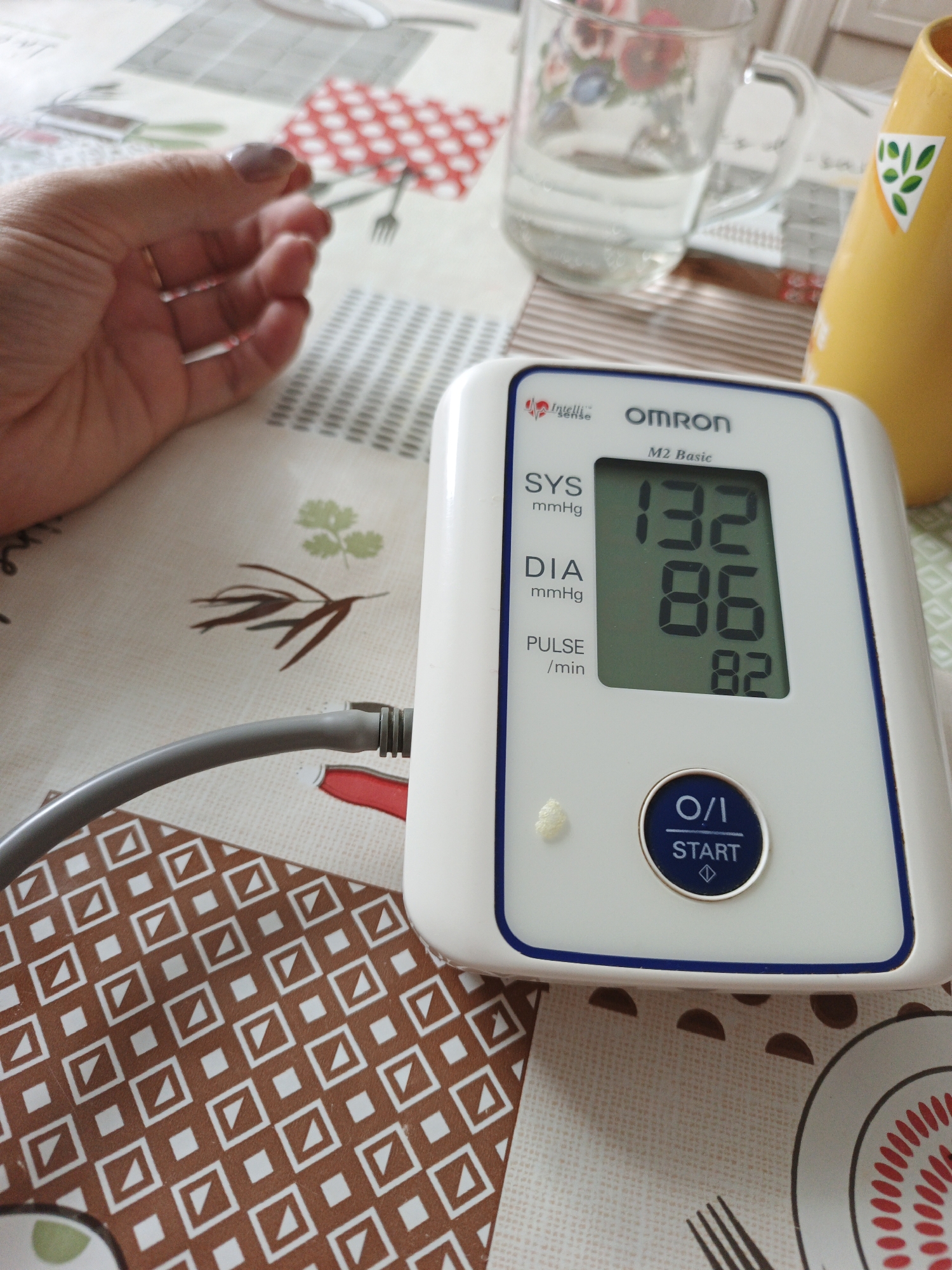 A digital Omron M2 Basic blood pressure monitor displays a systolic reading of 132 mmHg, a diastolic reading of 86 mmHg, and a pulse of 82 beats per minute. The device is situated on a table covered with a patterned tablecloth. To the left of the monitor, a person's hand rests on the table. In the background, a glass of water and a yellow vase are visible. The scene is indoors. The specific location in Medvedyovka, Russia is not identifiable from the image.