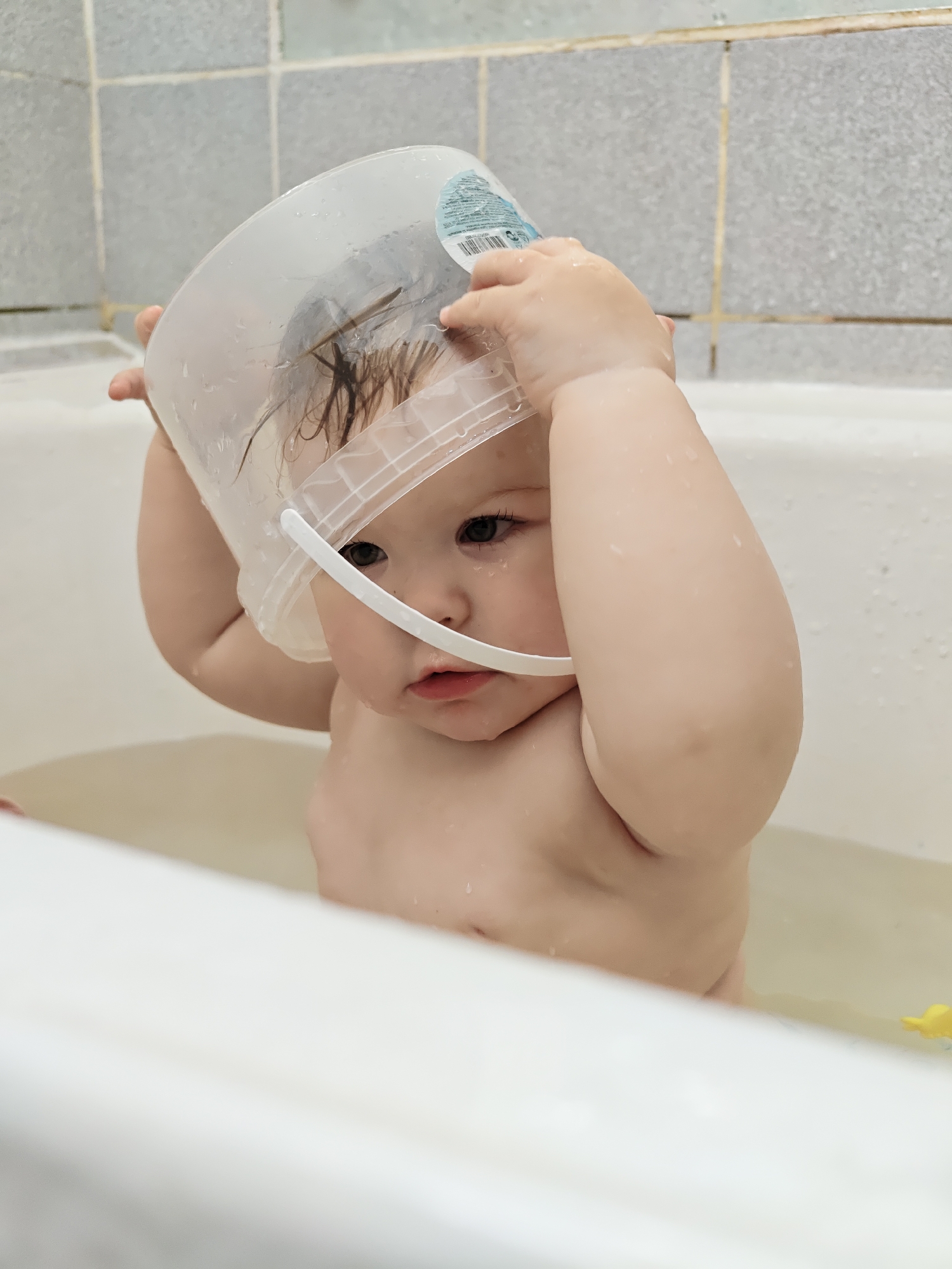 A baby is captured during bath time in a bathroom in Chaikovsky, Russia. The infant, with wet, dark hair and dark eyes, is seen from the chest up, partially submerged in shallow water within a white bathtub. The baby holds a clear plastic bucket with a white handle over its head, appearing to play with it or wear it. Water droplets are visible on the baby's skin and the translucent bucket. The baby's expression is neutral, appearing contemplative or curious as it interacts with the object.
In the background, a tiled wall with light grey or beige square tiles and darker grout lines is visible. The top edge of the white bathtub forms the foreground. A small, yellow bath toy is partially visible in the water to the right. A label on the clear bucket displays a barcode and some small, illegible text. The scene is well-lit, suggesting daytime.
