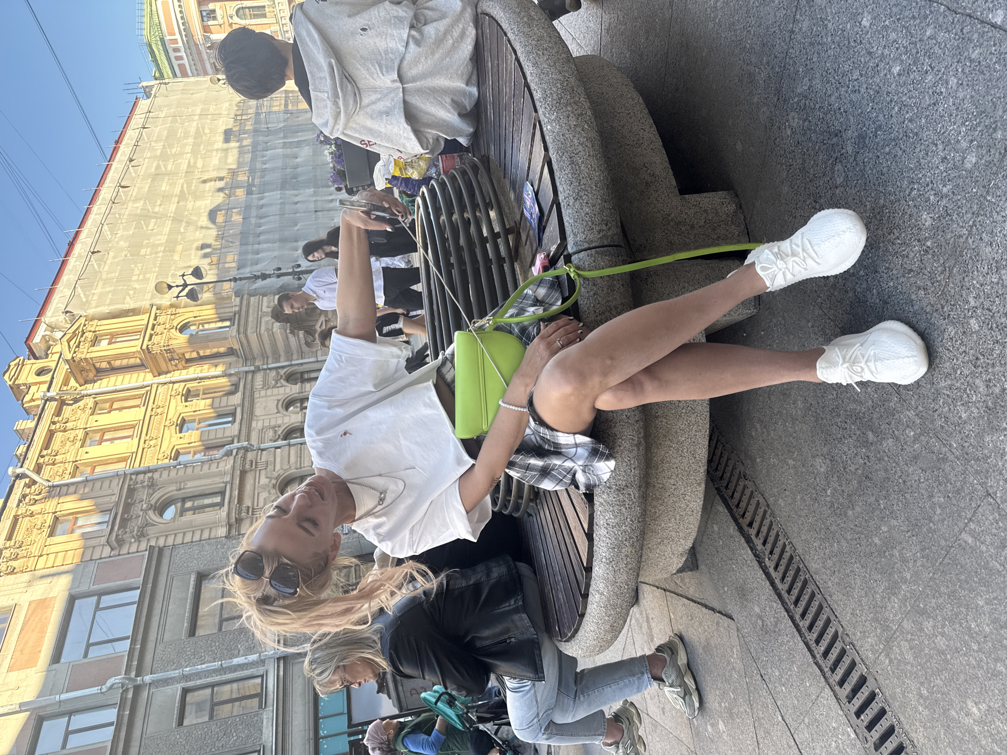 A blonde woman with sunglasses perched on her head leans back on a circular stone bench in St. Petersburg, Russia. She wears a white cropped t-shirt, a plaid skirt, and white sneakers, with a neon green purse slung over her shoulder. The bench is made of stone and wood, with a metal grate in the center. Behind her, other people are visible, including a woman in a black leather jacket and jeans sitting on the same bench to her left, and a person in a grey hoodie to her right. In the background, buildings with ornate facades line a sun-drenched street. The overall scene depicts a casual moment in an urban setting.