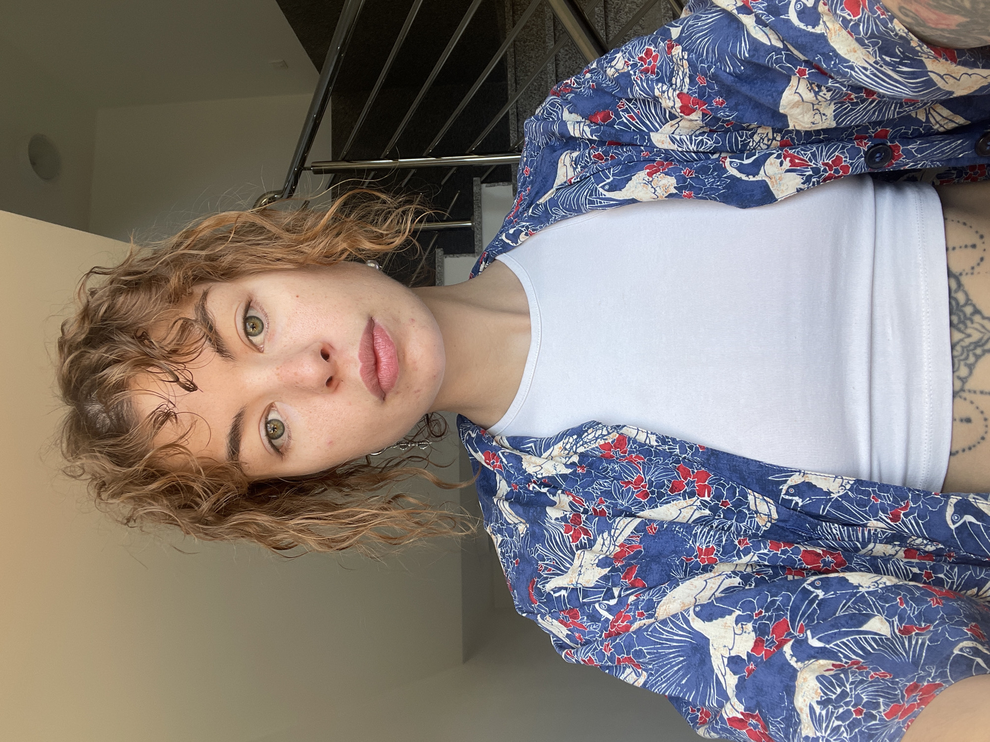 A young person with light brown, curly hair and fair skin is shown indoors, wearing a white top and a blue button-down shirt with a white bird and red flower pattern. The background shows a metal staircase and a light beige wall. The location is possibly in Boreti, Montenegro, based on the surrounding details.