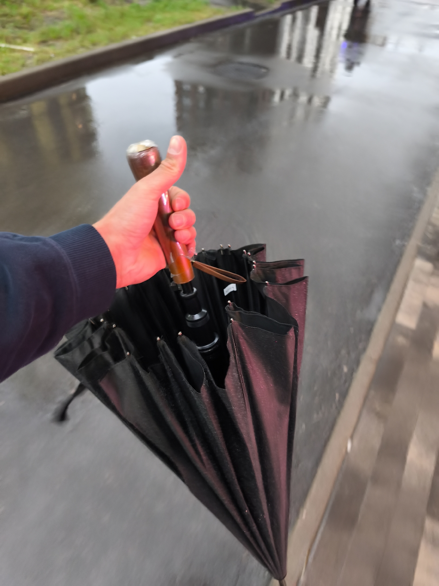 A person holds a closed black umbrella with a brown wooden handle on a wet city street, likely in Moscow, Russia. The person, wearing a dark blue long-sleeved shirt, has their hand gripping the umbrella handle. The street surface is reflective, indicating recent rain, and reflects nearby buildings. In the background, there is a hint of green vegetation along the edge of the sidewalk. The sidewalk is made of light and brown bricks. The perspective is first-person, giving the viewer the sense of holding the umbrella.
