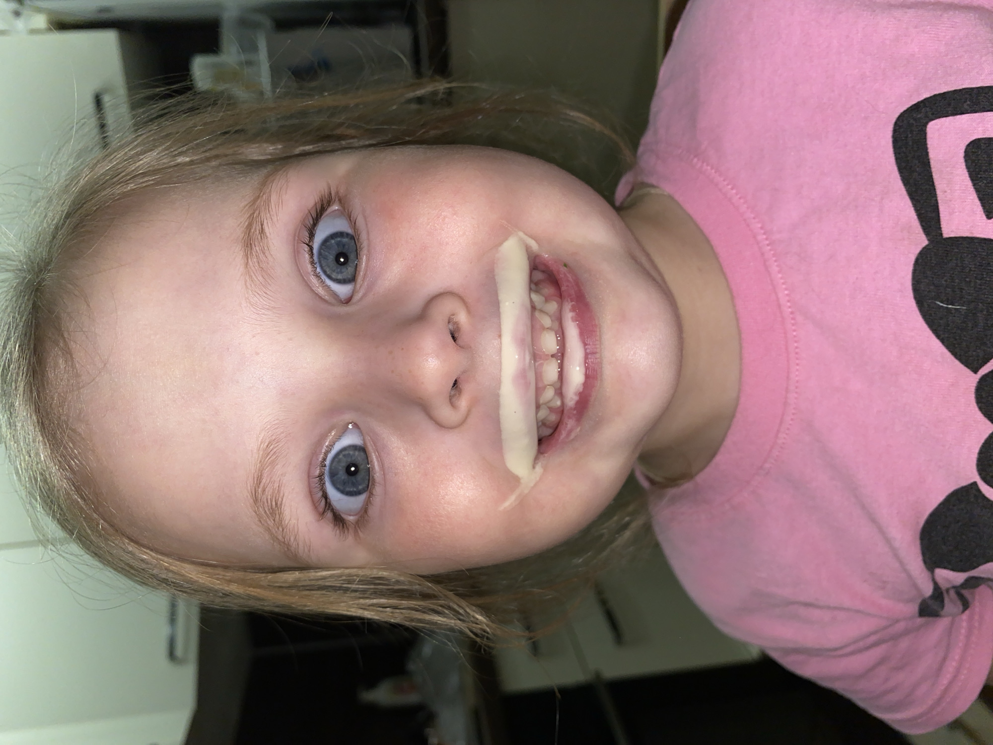 An indoor, close-up portrait shows a young child, likely a girl, looking directly at the camera with wide, blue eyes. Her light blonde hair frames her face. She is wearing a pink t-shirt with a dark, indistinct graphic design visible on her right shoulder area. A light yellowish, pasty substance, possibly food like mashed banana or yogurt, is smeared from the corner of her mouth across her cheek. Her mouth is slightly open, revealing some teeth behind the substance. The background is blurred, showing hints of an indoor setting with what appears to be light-colored cabinetry or furniture. The lighting suggests an indoor shot, with no specific cues for the time of day. The child's expression conveys a candid, possibly playful or surprised moment, captured during a messy activity. No discernible text is visible in the image.