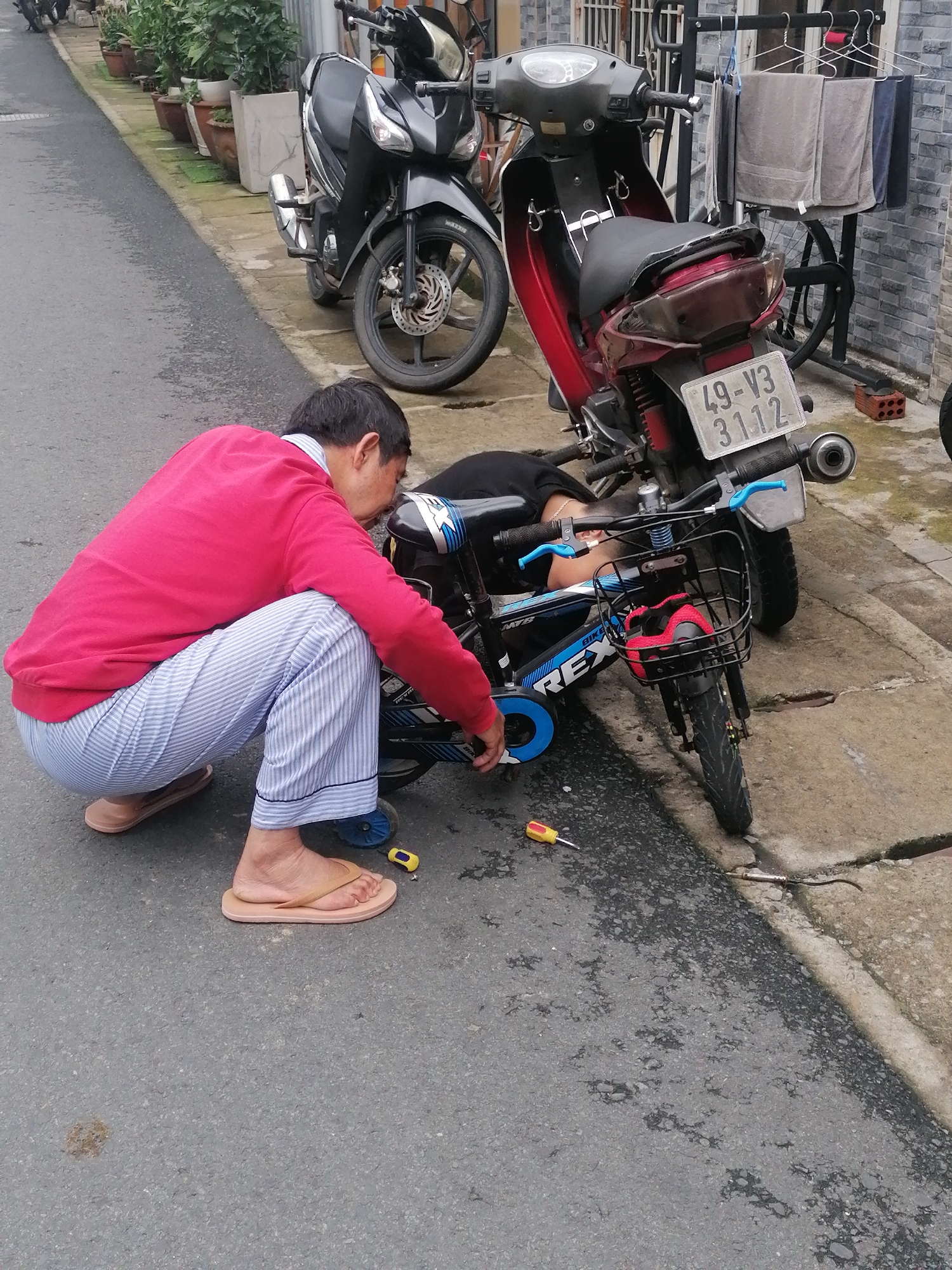 A man in a red shirt and blue and white striped pants is crouching on a road, apparently repairing a small blue and black bicycle. A child lies on the ground beside the bicycle. A red and black motorcycle is parked nearby, with a license plate reading "49-V3 3112." Another black motorcycle is parked further down the road, and potted plants line the sidewalk. A clothes-drying rack with clothes hanging on it stands against a building in the background. A yellow screwdriver lies on the road next to the man. The location is Da Lat, Vietnam.