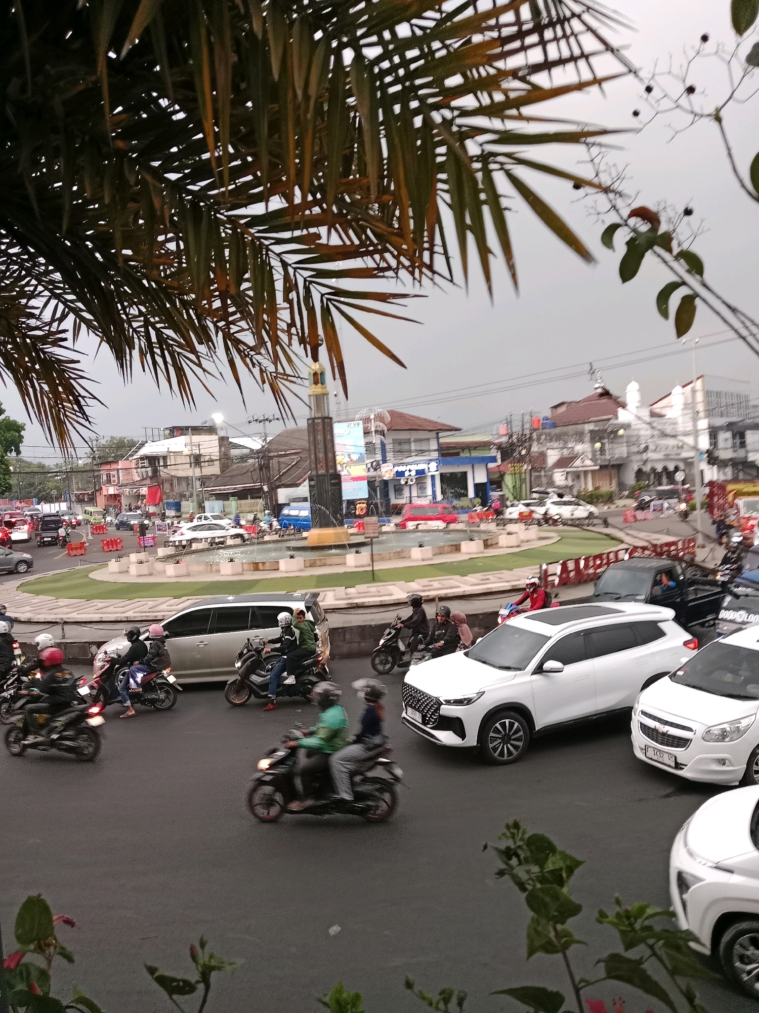 The image depicts a traffic scene at a roundabout in Cianjur, Indonesia. Palm fronds in the foreground partially obscure the view of the roundabout, which features a monument at its center and a fountain. Several vehicles, including cars and numerous motorcycles, are navigating the roundabout. People on motorcycles are wearing helmets and casual attire. Buildings and shops are visible in the background, with one establishment displaying a sign that appears to say "TOKO ROTI" (bakery). The sky is overcast, suggesting potentially humid weather. The overall impression is one of a busy urban intersection in Indonesia.