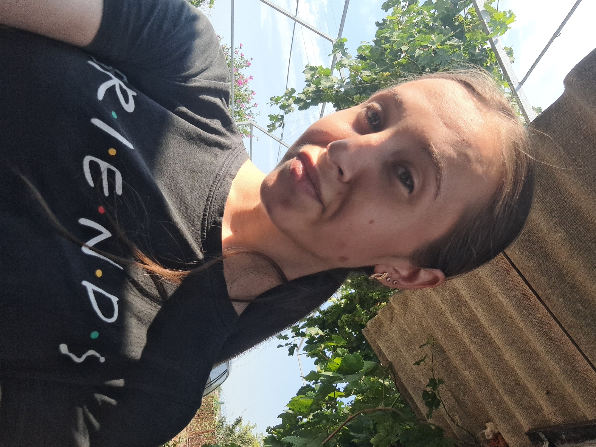A young woman is taking a selfie outdoors. She is wearing a black t-shirt with the "Friends" logo on it. She has brown hair pulled back in a ponytail and is wearing earrings. The background shows green foliage, a clear sky, and a corrugated roof. The location is Vypasne, Ukraine.