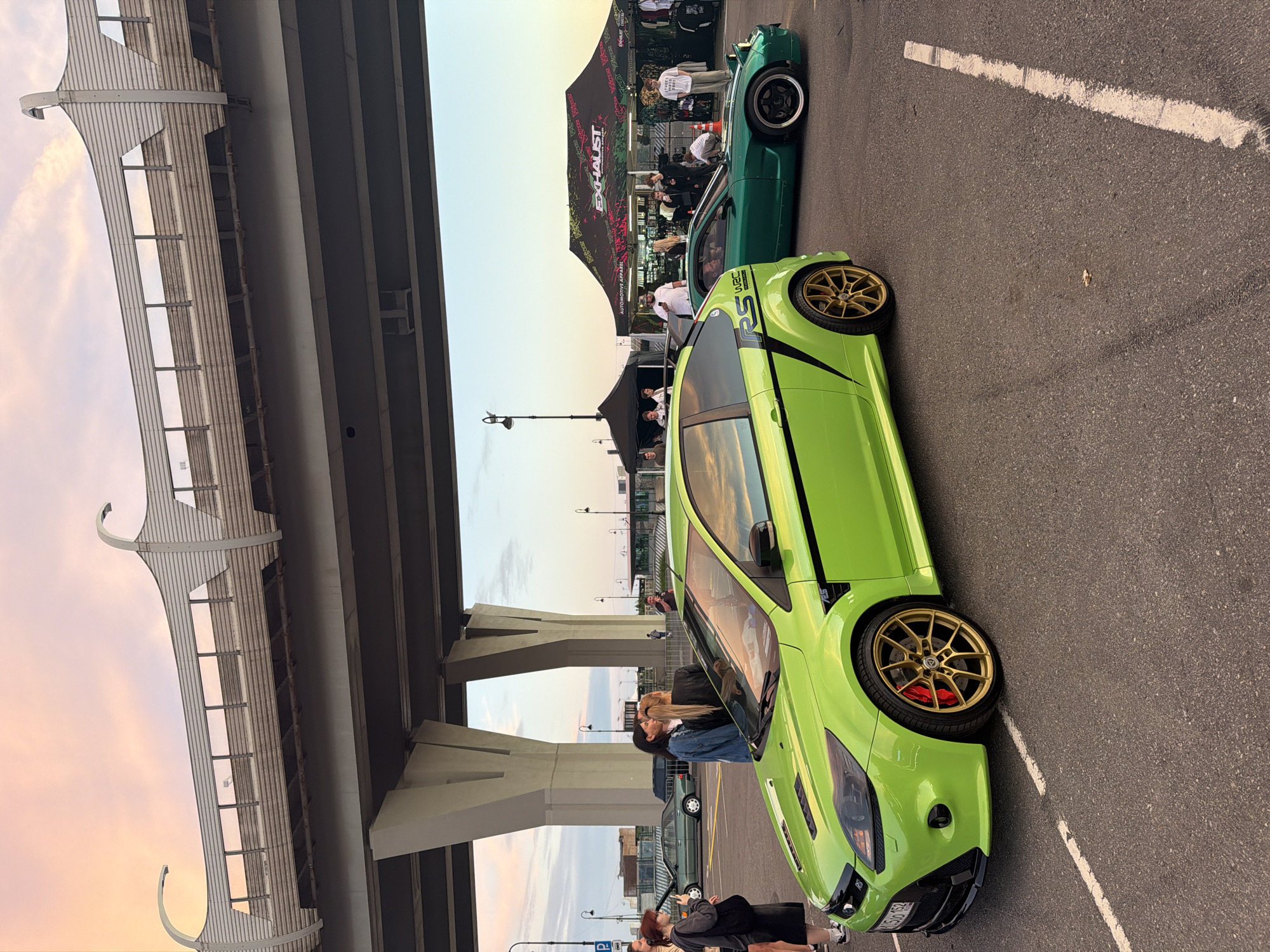 The image shows an outdoor car show under an overpass. In the foreground, a lime green Ford Focus RS is parked. The car has gold rims and red brake calipers. To the right of the green car is a dark green Mazda RX-7. Behind the cars, a black tent and a large white canopy with the "EXHAUST" logo are set up. Several people are milling around the cars and tents. In the background, a vintage green sedan is parked. The sky is a soft pink and orange, suggesting it is sunset. The ground is asphalt, with white painted parking lines. The overpass above is a prominent concrete structure with supporting columns.