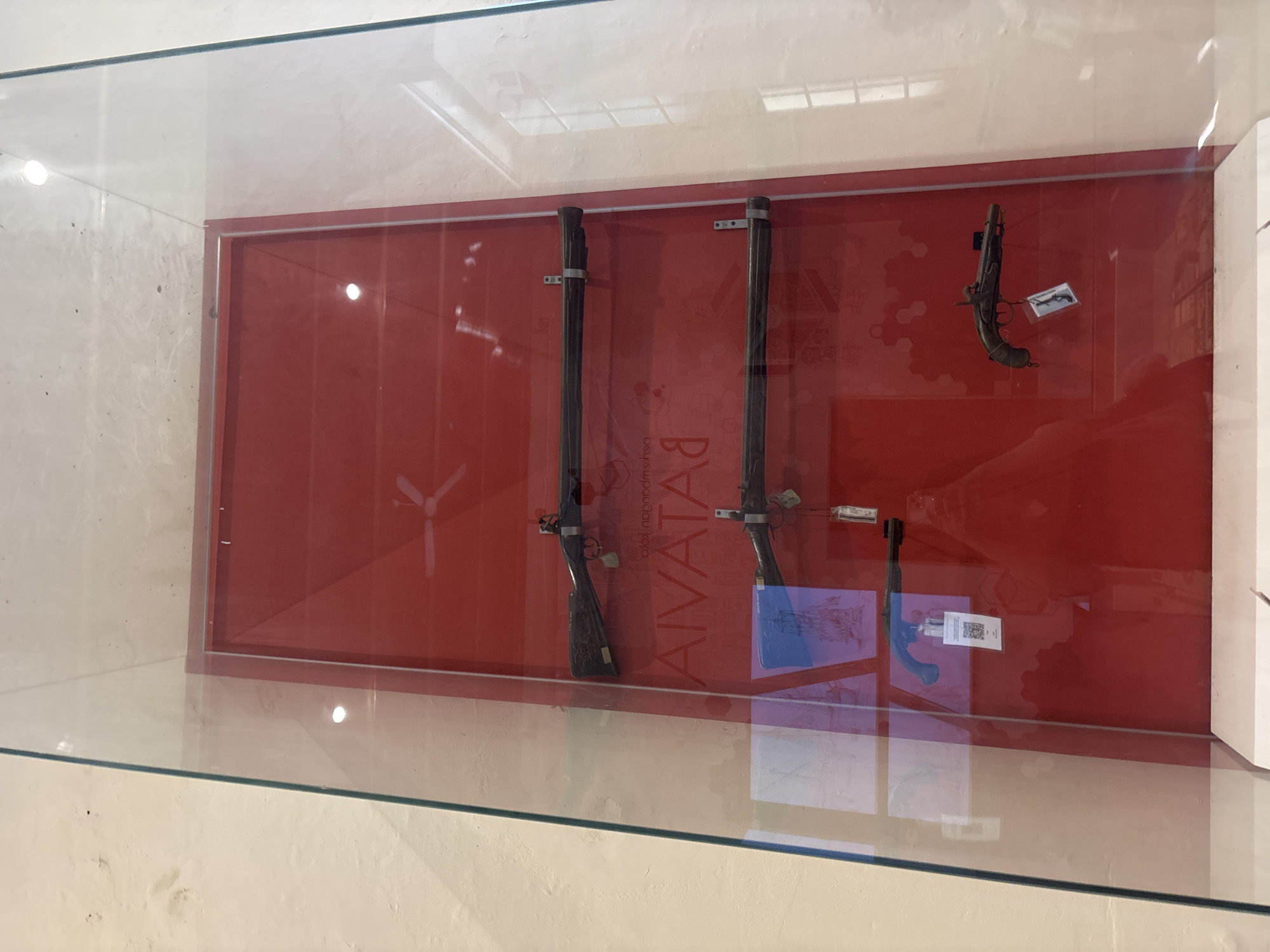 The image depicts a museum display case featuring antique firearms, specifically three rifles and two pistols, mounted on a red background. The display case is made of glass and is situated against a light-colored wall. Visible in the background are the outlines of ceiling fans and window panes, suggesting an indoor exhibition space. The text "AIVATA" is prominently displayed on the red background behind the firearms. This exhibit is located in the Museum Fatahillah, also known as the Jakarta History Museum, situated in the Old Town of West Jakarta, Indonesia.