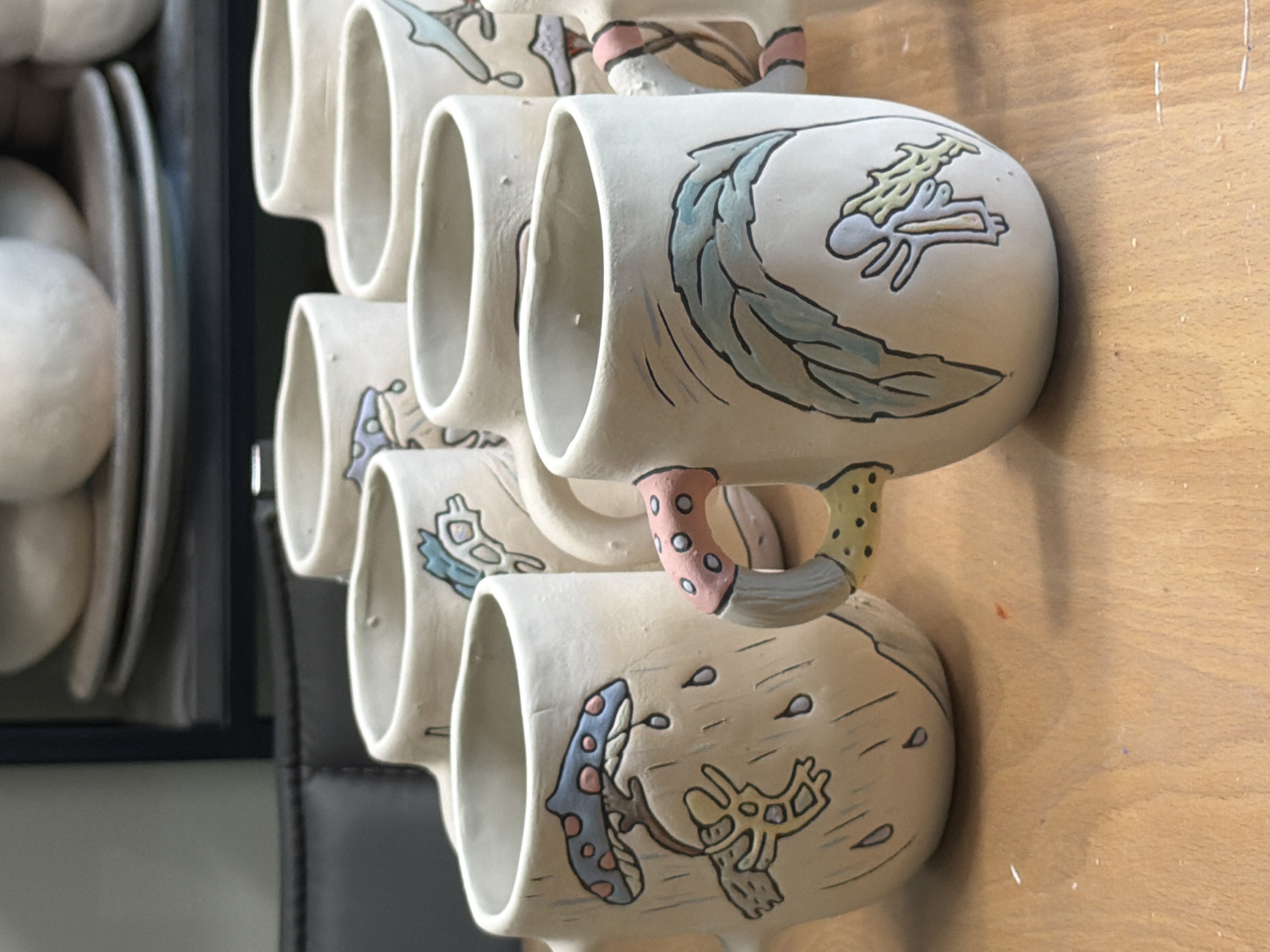 The image shows a close-up, eye-level, medium shot of several handmade ceramic mugs on a wooden surface. The mugs are off-white with hand-painted illustrations. The mug in the foreground on the left features a blue and pink spotted mushroom with falling water droplets and a yellow, abstract figure. The mug to its right has a handle with pink and white spots and a yellow base with green spots. The mug in the right foreground displays a green, leafy design and a small, light blue figure with yellow accents. In the background, other mugs and stacks of ceramic plates are visible, as well as a dark leather-like surface and shelving unit. The scene appears to be indoors, possibly in an art studio or craft workshop. The location in the city of Askat, Russia is not discernible from the image.