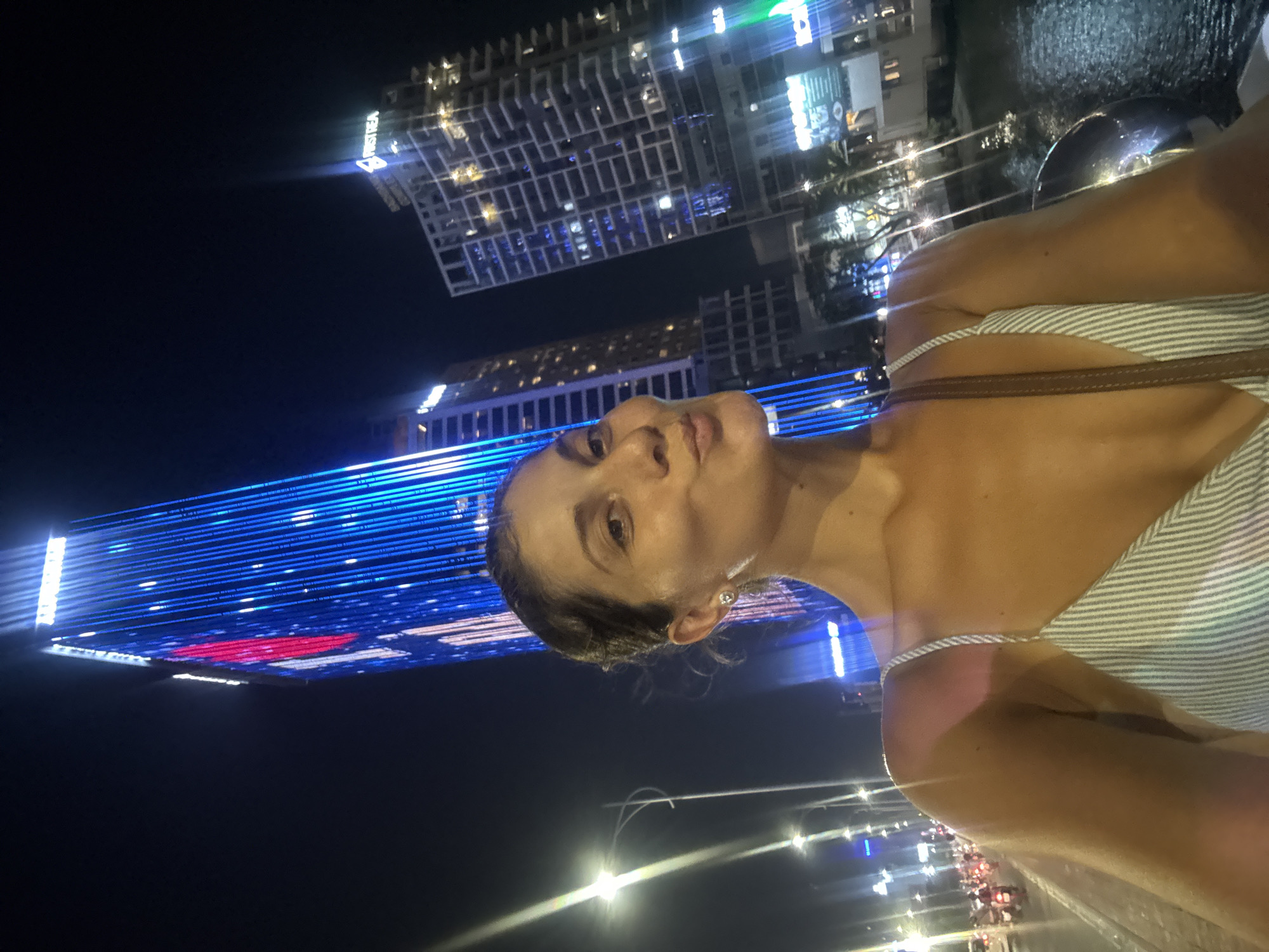 A nighttime shot of a woman standing in Đà Nẵng, Vietnam, with the city's skyline as a backdrop. She has her head tilted back. Behind her are illuminated buildings and city lights. One building has a large display of blue lights with a red section on it. Other buildings are lit with white and yellow lights. To the side, there is the Han River. The woman is wearing a striped top.