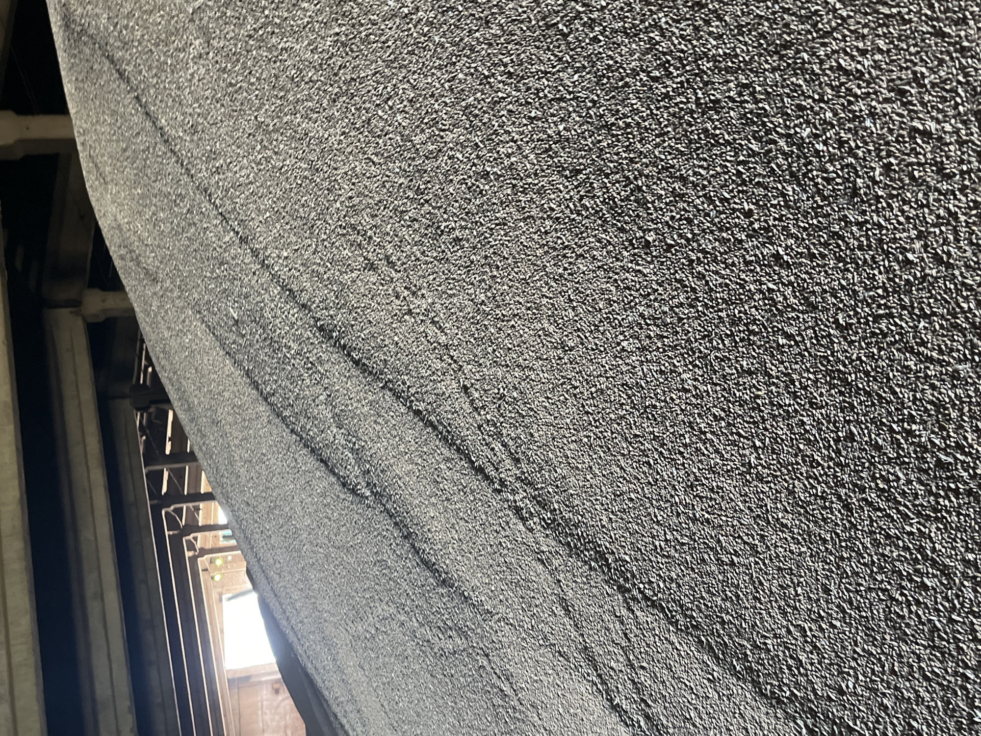 The image shows a close-up view of the interior of a structure, possibly a grain elevator, in Unknown, Unknown. A gray, textured wall fills most of the frame. The wall appears to have a rough, grainy surface. To the left, there is a glimpse of a structural framework of metal beams and supports, leading towards an opening where light is visible. The lighting is dim, casting shadows within the framework and highlighting the texture of the wall.