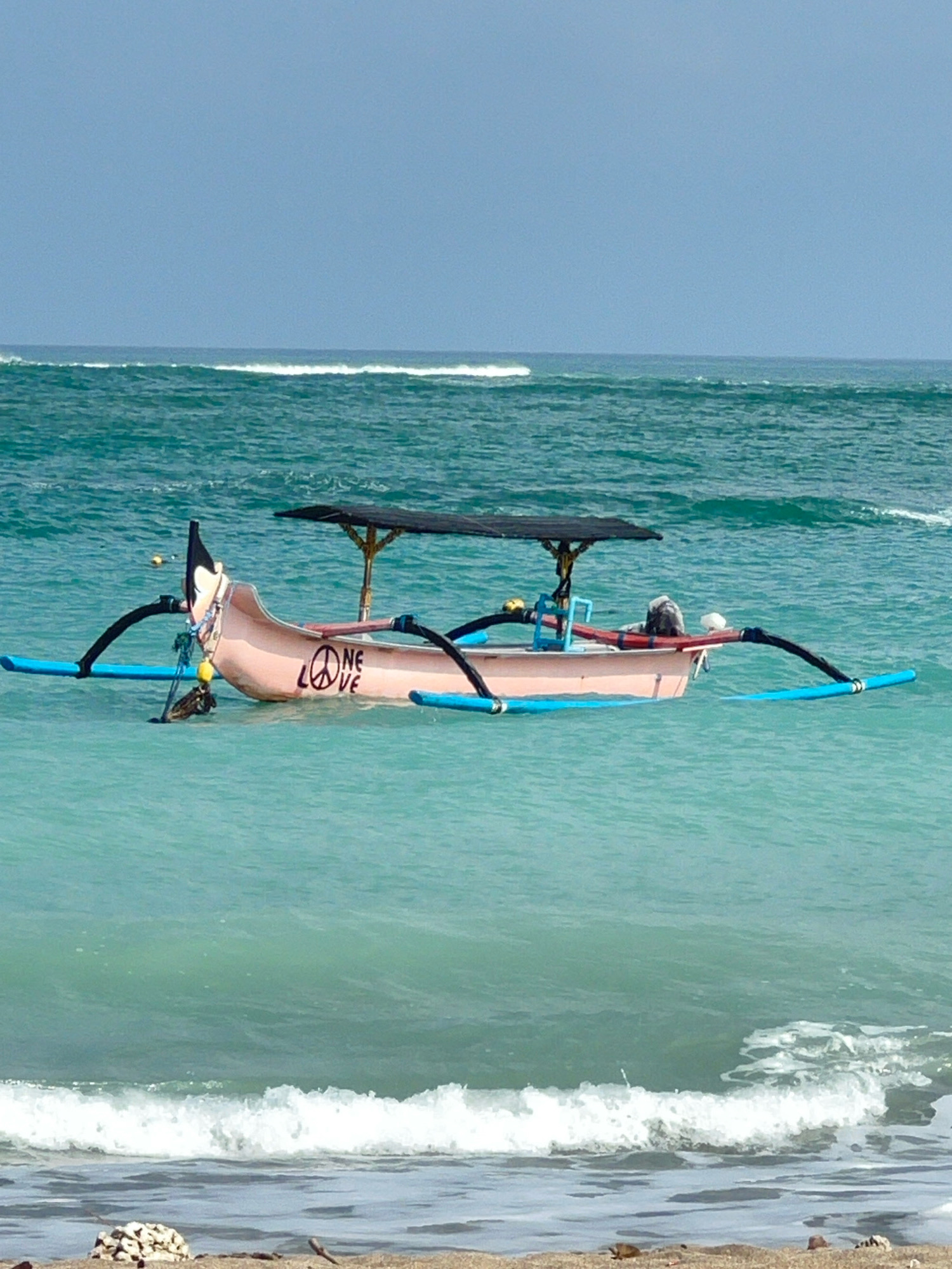 A single outrigger boat floats on turquoise water near the shore of Kuta, Indonesia. The boat is pink with black outrigger floats and a black canopy. A peace symbol and the words "ONE LOVE" are painted on the side of the boat. The sky is clear and blue, and small waves are breaking on the beach in the foreground. The scene is tranquil and serene, with the boat appearing to be at rest.