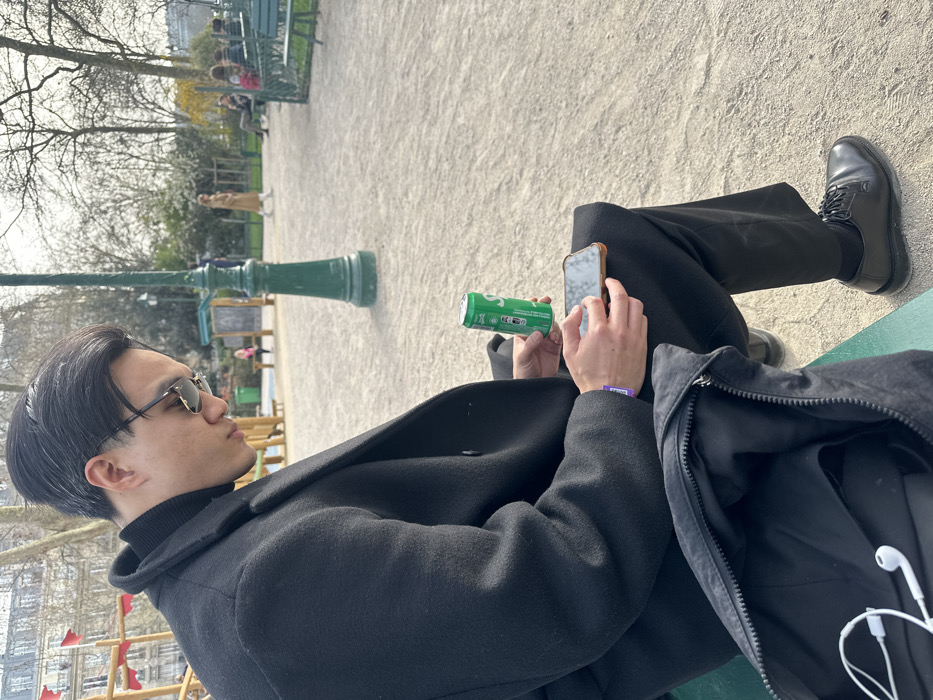 An adult male is seated on a green public bench in a Parisian park during daytime. He is wearing a black turtleneck, black overcoat, black trousers, and black leather shoes, with dark hair and sunglasses. His gaze is directed downwards at a smartphone held in his left hand. His right hand holds an open green Sprite can. A purple wristband is visible on his left wrist. An open black bag, containing white wired earbuds, rests beside him on the bench. The ground consists of light-colored gravel. In the middle ground, a tall, dark green lamppost stands. The background features sparse, deciduous trees, indicative of late winter or early spring, and other park visitors, including a woman in a long light-colored coat walking and several individuals seated on distant benches. A children's playground with a red structure is visible to the far left. Beyond the park's trees and features, classic Parisian buildings with light-colored stone facades are present, confirming the location as a public garden within Paris, France.