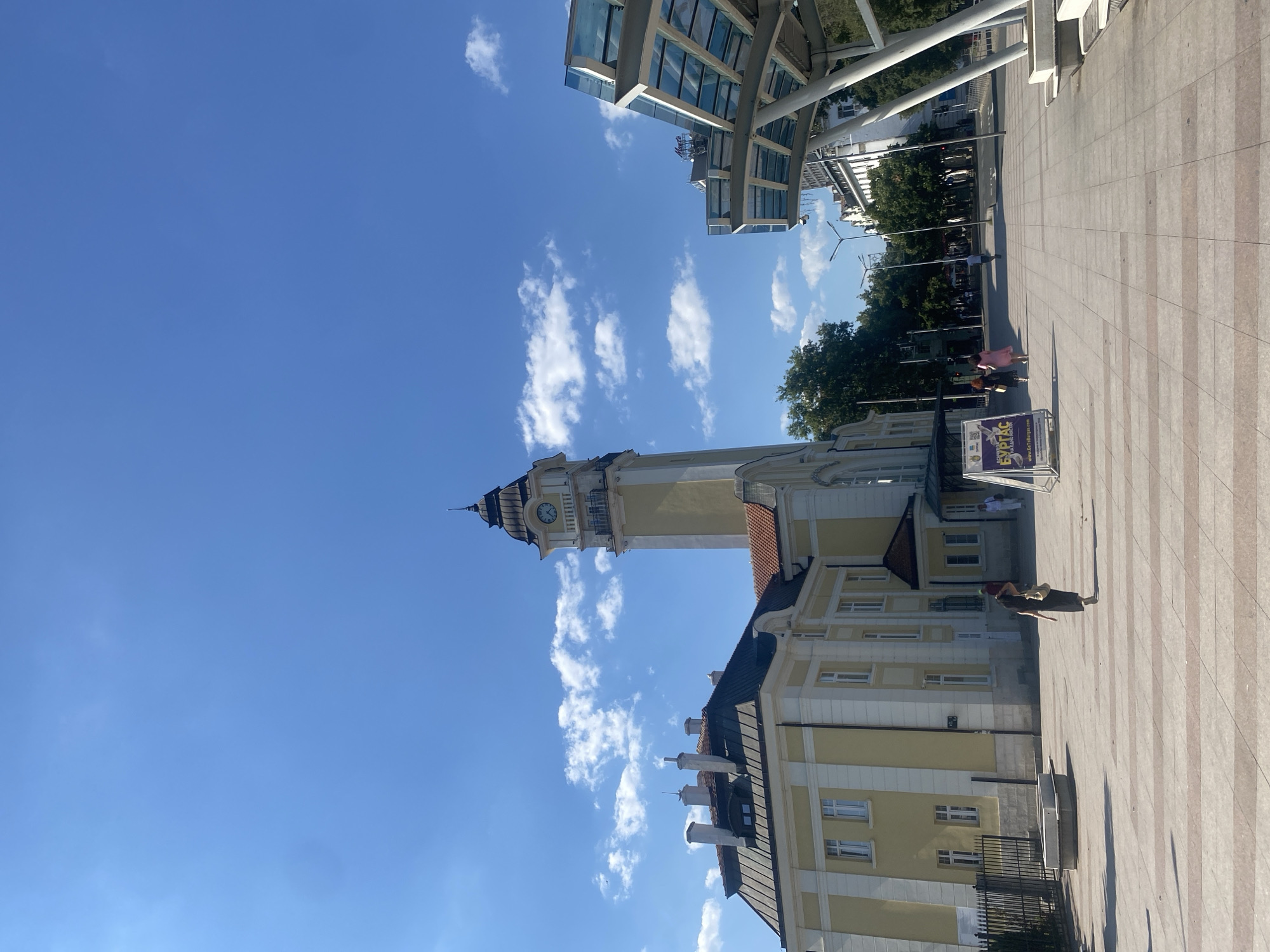 The image shows a wide, paved plaza in front of a yellow and white building with a tall clock tower. The building is a historic structure, possibly a town hall or administrative building, with classical architectural elements. A modern glass and metal structure, possibly a bus stop shelter, is visible to the right. The sky is blue with scattered white clouds. Several people are walking across the plaza. In the background, trees and other buildings line a street. A sign in Bulgarian is displayed on an easel in the foreground. The scene depicts a public space in the city of Burgas, Bulgaria, likely near the city center given the architectural style and public amenities.