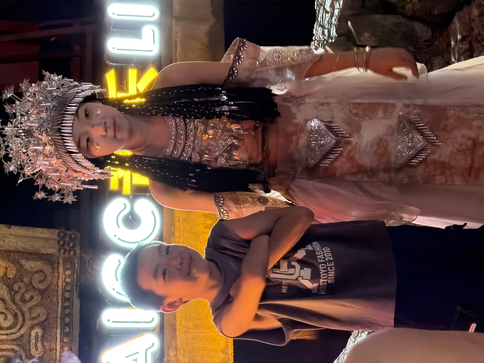 In Yuzhong District, China, a young boy and a young woman stand together outdoors at night. The boy, on the left, wears a grey t-shirt with the text "HOSTOYO FASHION SINCE 2010" and dark pants. His arms are crossed, and he is smiling. The young woman, on the right, is dressed in an elaborate costume featuring a silver headdress, intricate beadwork, and flowing fabric. She has dark hair styled in braids adorned with silver accents and glitter on her face. Behind them, large, illuminated neon signs with the letters "AIC" and "LI" glow in bright white and yellow. The background includes decorative stonework and a rocky texture, suggesting an outdoor entertainment area or attraction.