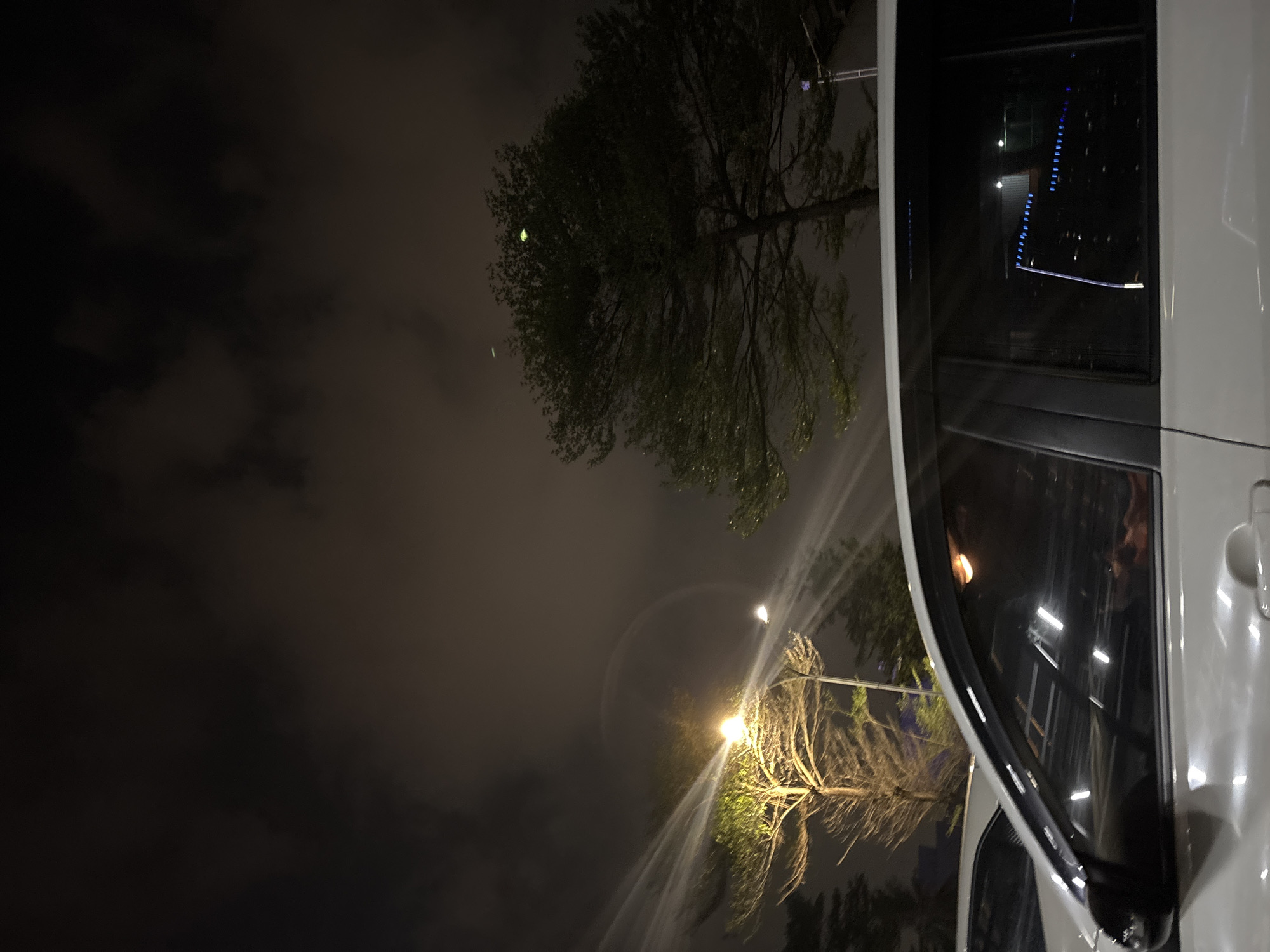 This is a night-time, eye-level, medium shot photograph taken in North Jakarta, Indonesia. The image features the side of a white car in the foreground, its windows reflecting streetlights and possibly buildings. Beyond the car, two trees are visible. The tree on the left is illuminated by a streetlamp, highlighting its branches and leaves. The tree on the right is darker and more densely foliated. The sky is dark with scattered clouds, and a light beam from a streetlamp cuts across the left side of the frame, creating a halo effect around a circular light source. The overall atmosphere is dim and moody, typical of a city at night.