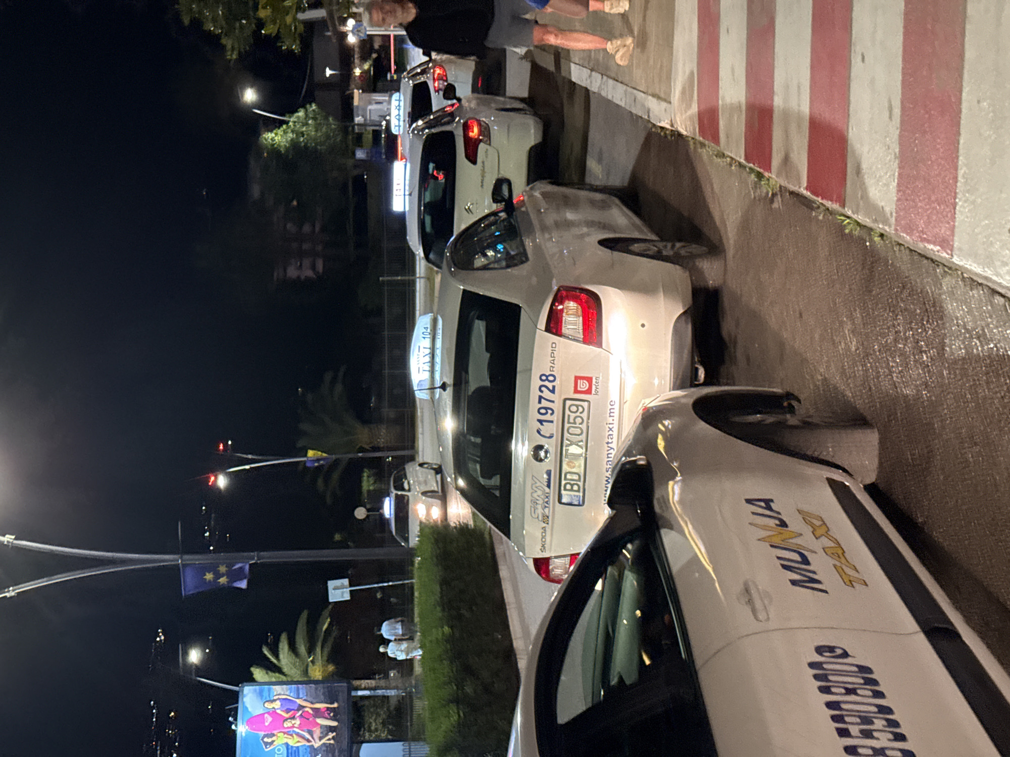 The image captures a nighttime street scene in Budva, Montenegro, focusing on a line of white taxis. The taxis are parked along a curb with red and white striped pedestrian crossing markings. In the foreground, a white taxi with "MUNJA TAXI" and the phone number "8 590 800" is partially visible. Behind it, another white taxi displays "SANY TAXI", a phone number "19728", and a license plate "BD TX 059". Further back, additional white taxis with illuminated "TAXI" signs are visible. The background shows a dimly lit urban environment with trees, lampposts, and distant buildings. Two individuals, a man and a woman, are walking on the sidewalk in the background. The overall impression is of a taxi stand operating at night in a coastal city.