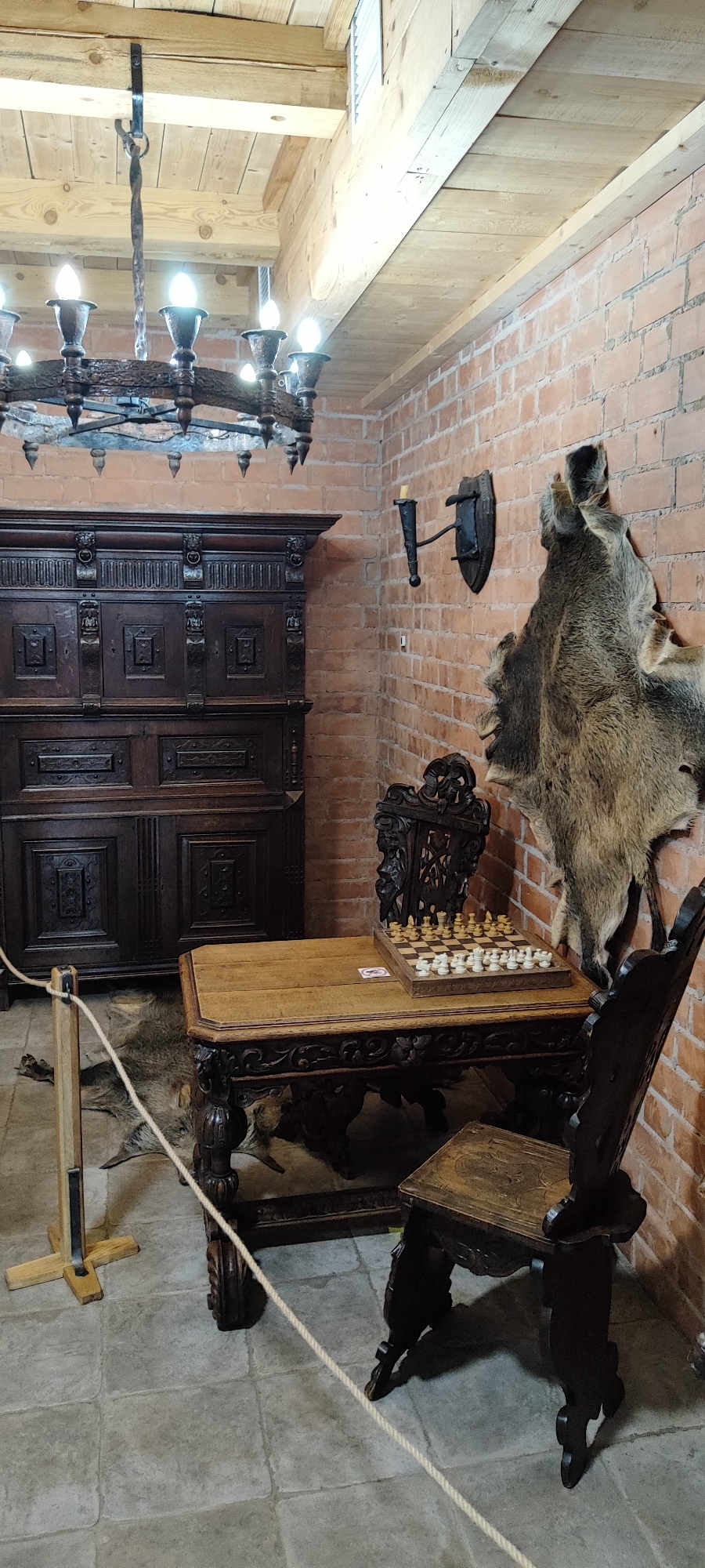 The image depicts an interior scene that appears to be part of a historical exhibition or museum, possibly within the Lida Castle in Lida, Belarus. The setting is characterized by rustic and medieval aesthetics.
In the background, a brick wall forms the right side of the frame. Mounted on the wall is a torch sconce, and a large animal pelt, possibly of a wild boar, is hung to the right of the sconce. To the left of the brick wall stands a tall, dark wooden cabinet with intricate carvings. Above the cabinet, a rustic chandelier with multiple lights hangs from a wooden ceiling structure.
In the foreground, a wooden table is positioned with a chessboard set up on it, complete with chess pieces. A decorative wooden chair with a carved back is placed behind the table. To the right of the table, another dark wooden chair is visible, partially in shadow. A rope barrier is set up in front of the table, suggesting that visitors are meant to observe the exhibit from a distance. A wolf pelt is visible on the stone floor in front of the cabinet and under the table. The floor is made of large, square stone tiles. The lighting in the scene is a mix of artificial light from the chandelier and possibly natural light from an unseen source, casting shadows that enhance the atmospheric quality of the display.